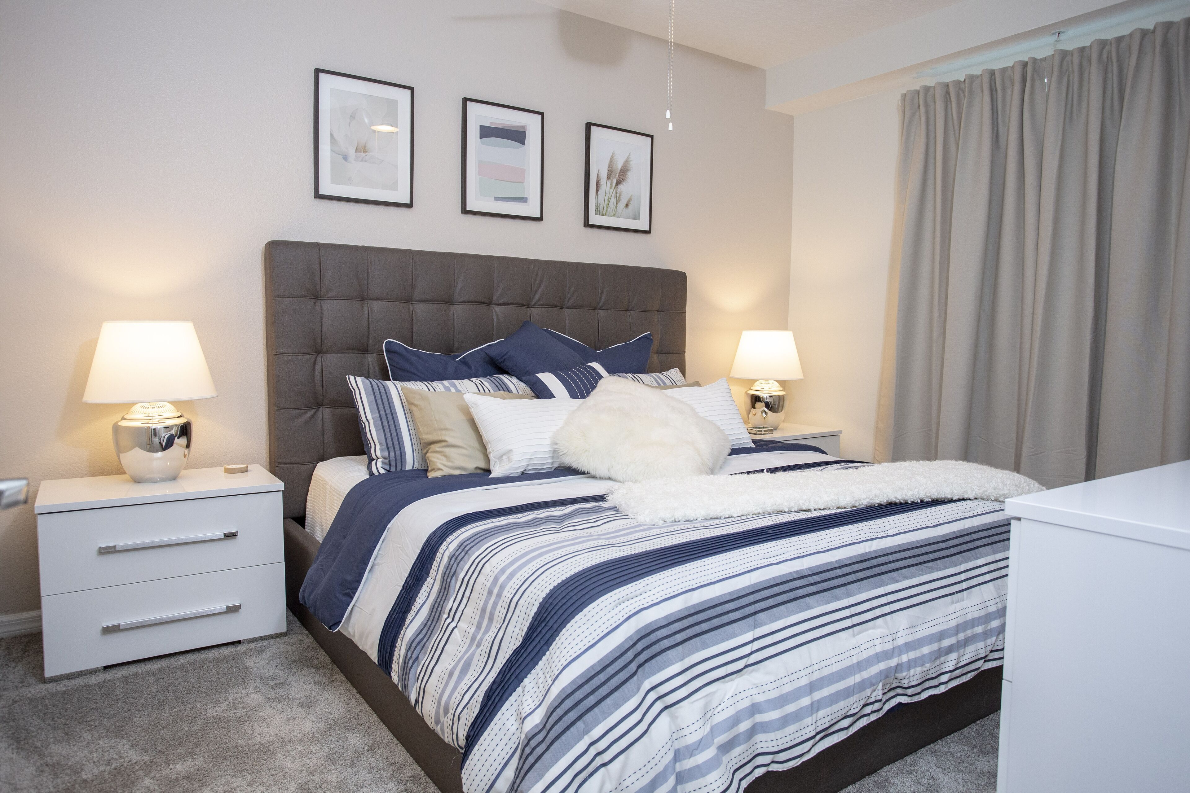 Une chambre à coucher avec une tête de lit capitonnée, une couvre-lit à rayures, deux tables de chevet avec des lampes et une grande fenêtre avec des rideaux.