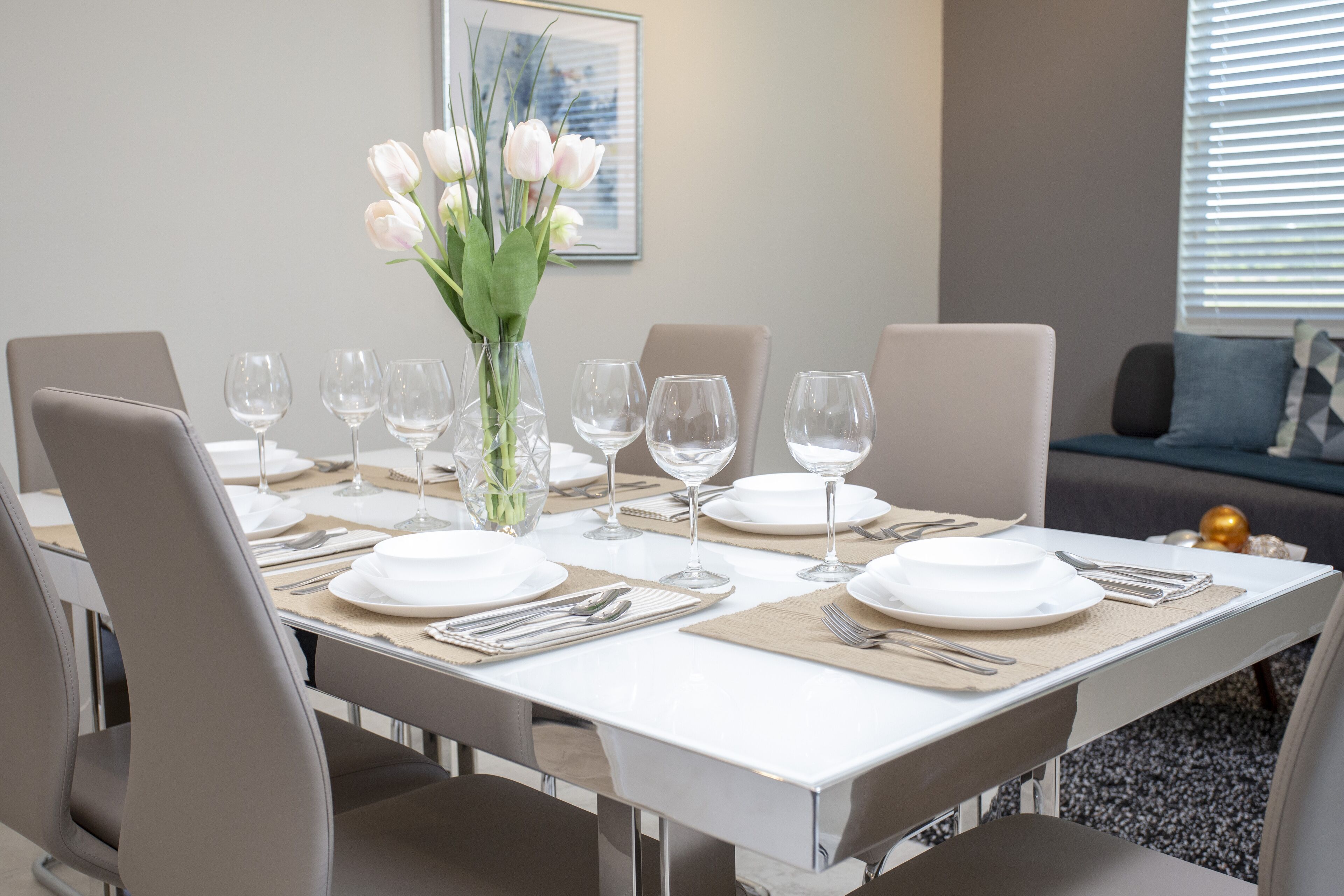 Comedor con una mesa blanca dispuesta para seis personas, rodeada de sillas grises. Un jarrón con flores blancas está sobre la mesa.