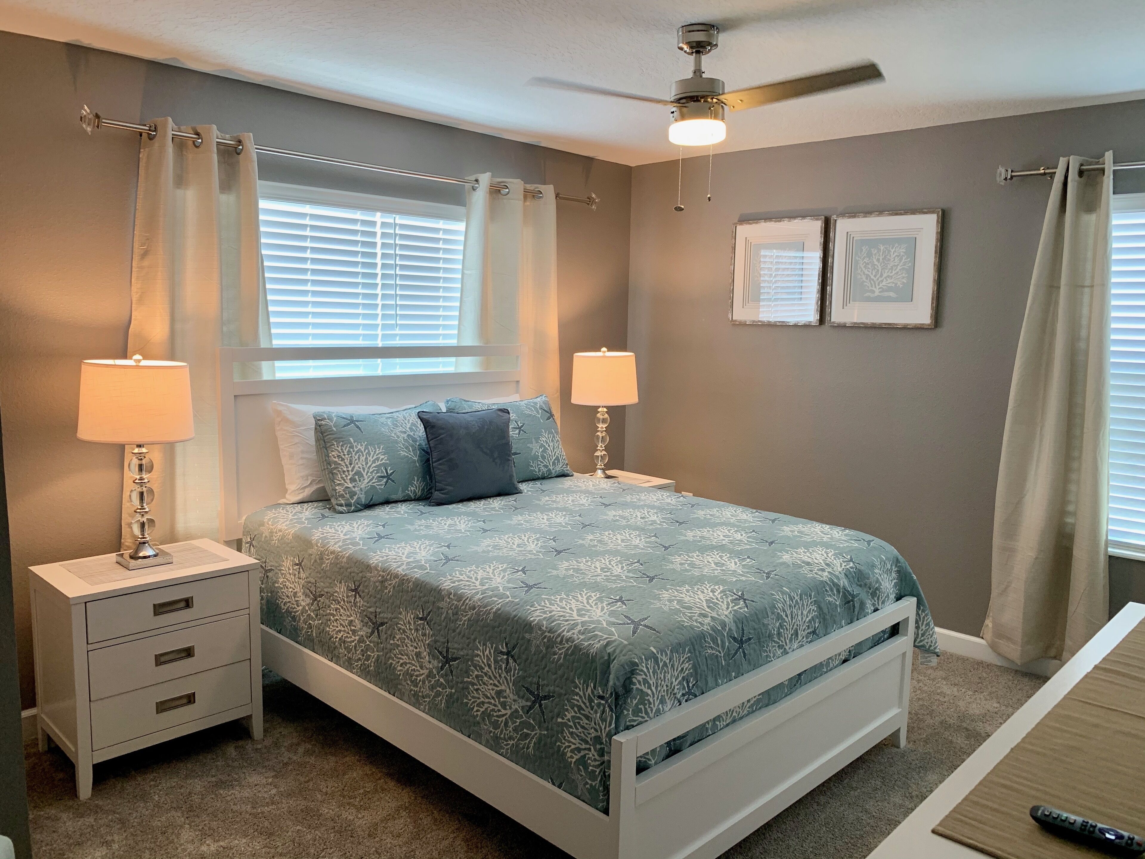 A bedroom with a bed, two bedside tables with lamps, a ceiling fan, and two framed pictures on the wall.