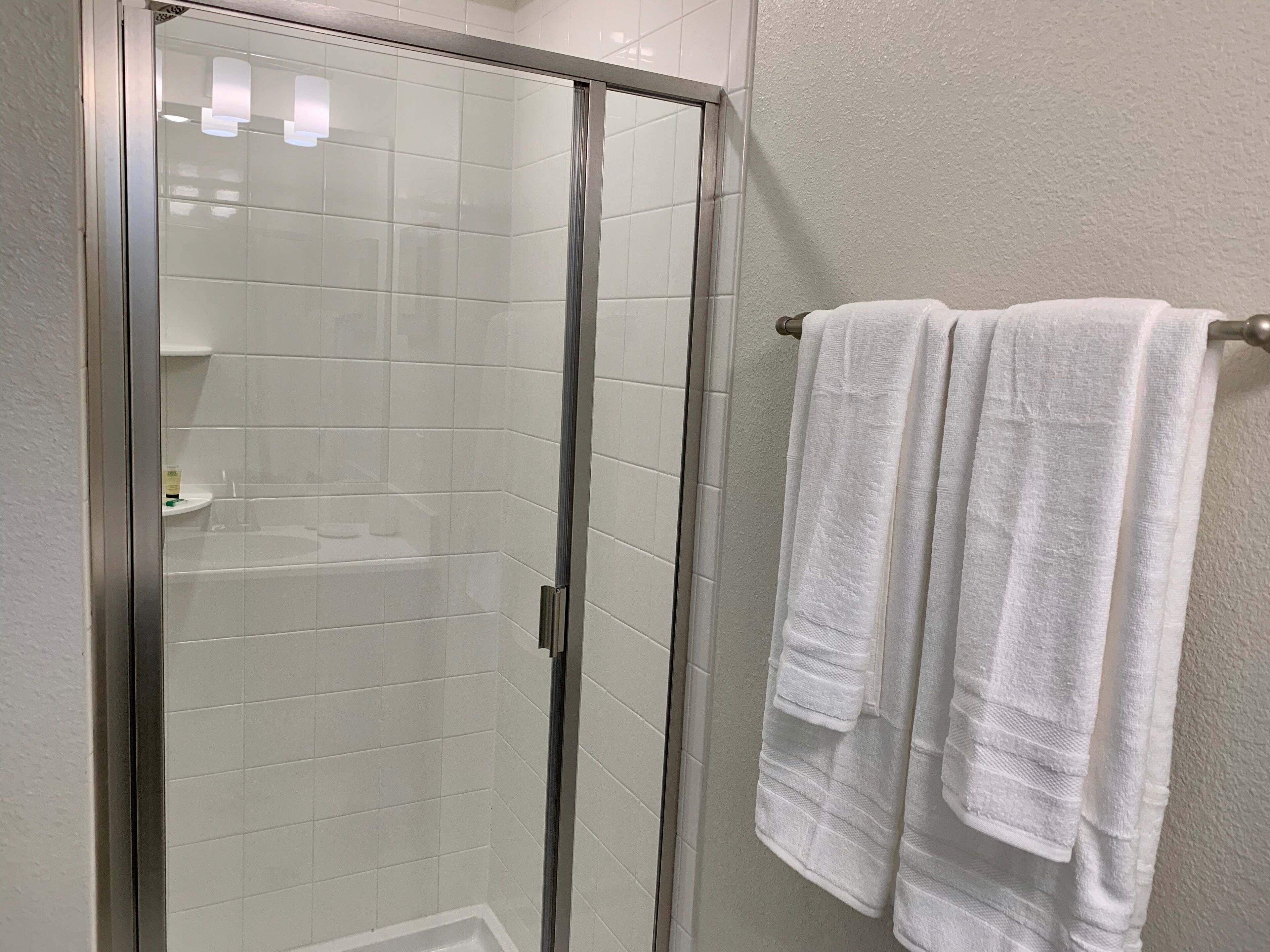 A glass-enclosed shower with white tiles, a towel rack with white towels, and a small shelf with toiletries.
