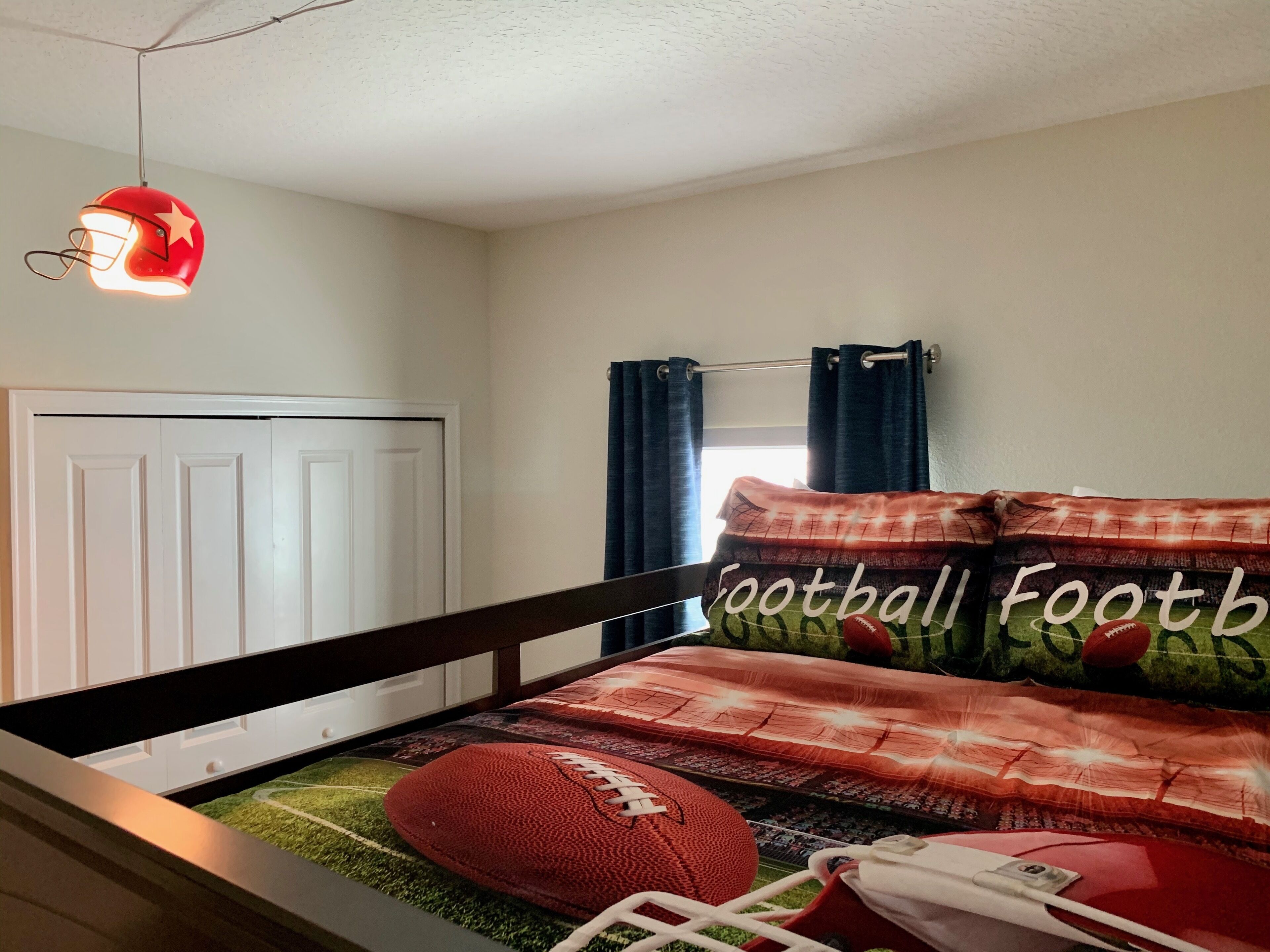 A bedroom with a football-themed bedspread, a red helmet light fixture, and a window with curtains.