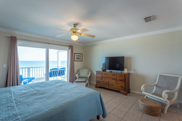 A bedroom with a bed, a TV, a dresser, and a chair. There is a balcony with a view of the ocean.