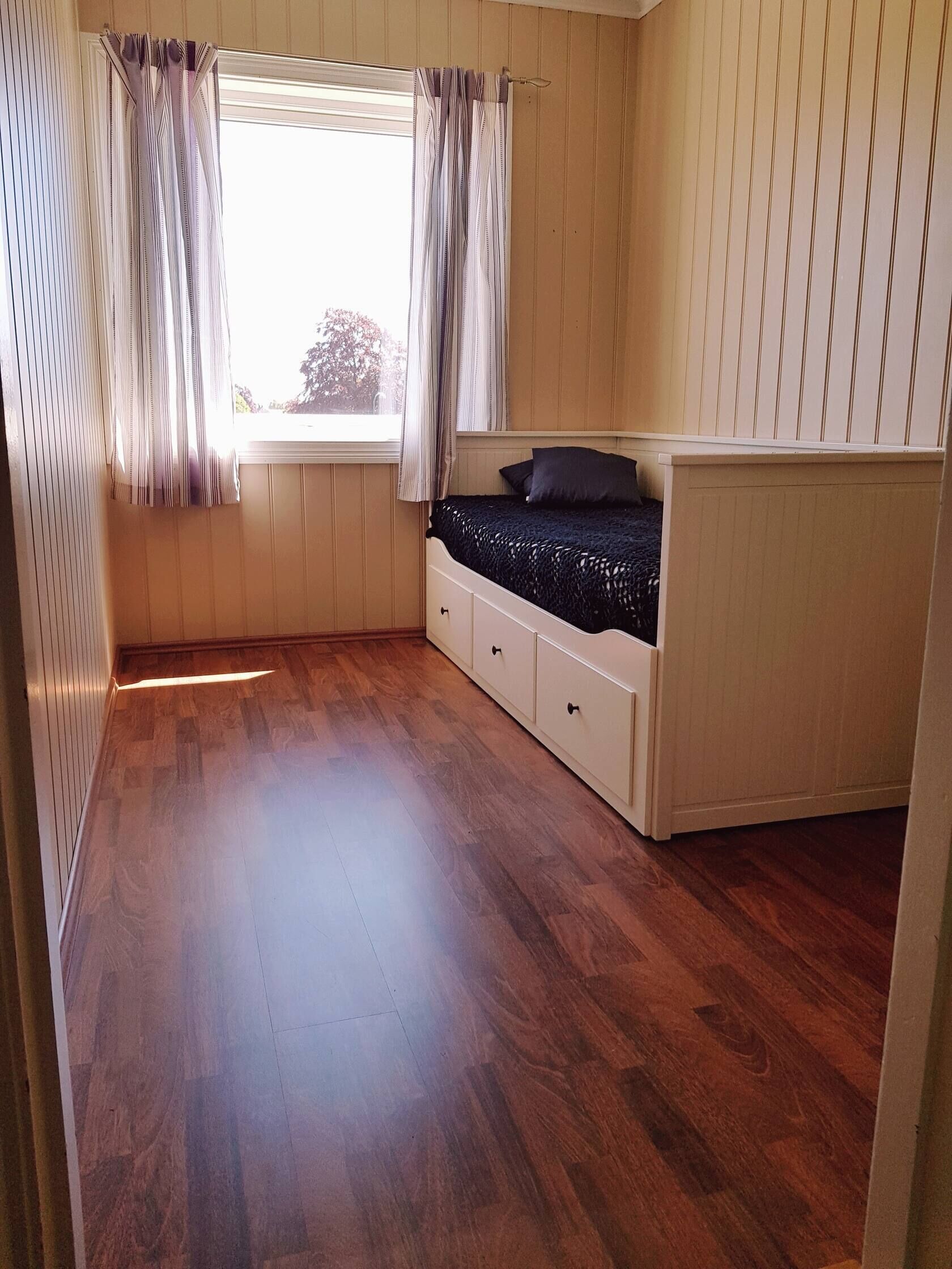 A bedroom with a wooden floor, a bed with a black cover, a window with curtains, and a radiator.
