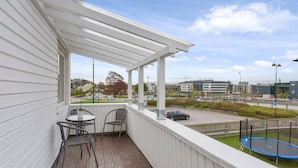 A balcony with a white railing, a small table with two chairs, and a view of a cityscape with buildings and a road.