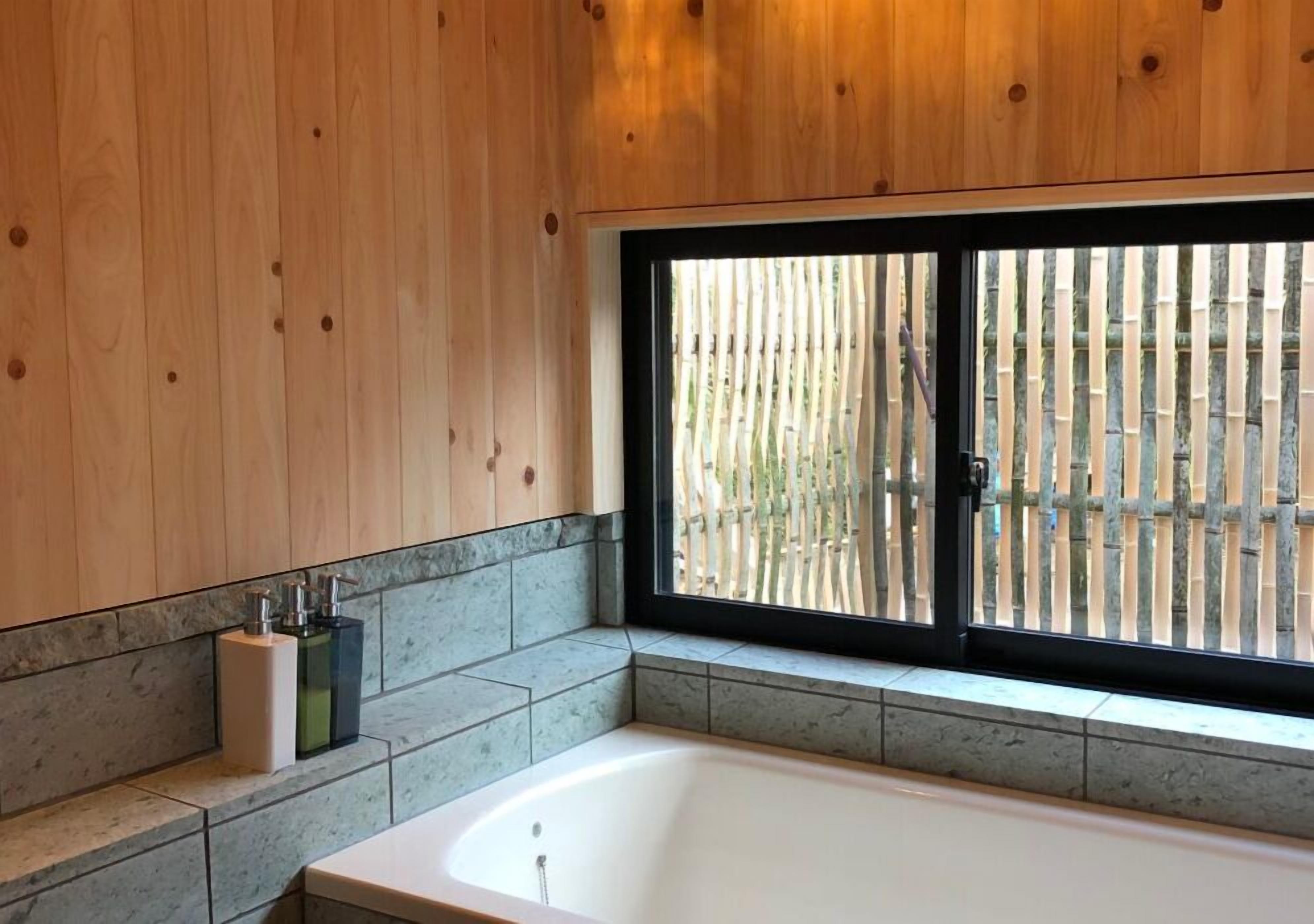 Un baño moderno con una bañera independiente, una ventana que ofrece vistas a una valla de bambú y una pared revestida de mármol.