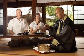 A traditional Japanese setting with tatami flooring, a low table, and a person in a black robe serving tea. - Temple Hotel Daitaiji (Nachikatsuura)
