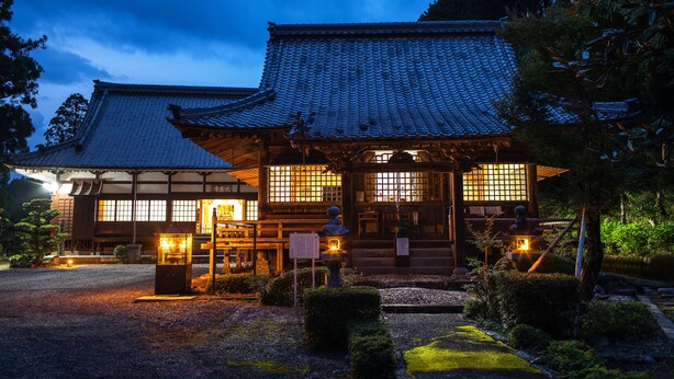 Un bâtiment japonais traditionnel, doté d’un toit en tuiles et d’une structure en bois, entouré d’un chemin de gravier et de verdure.