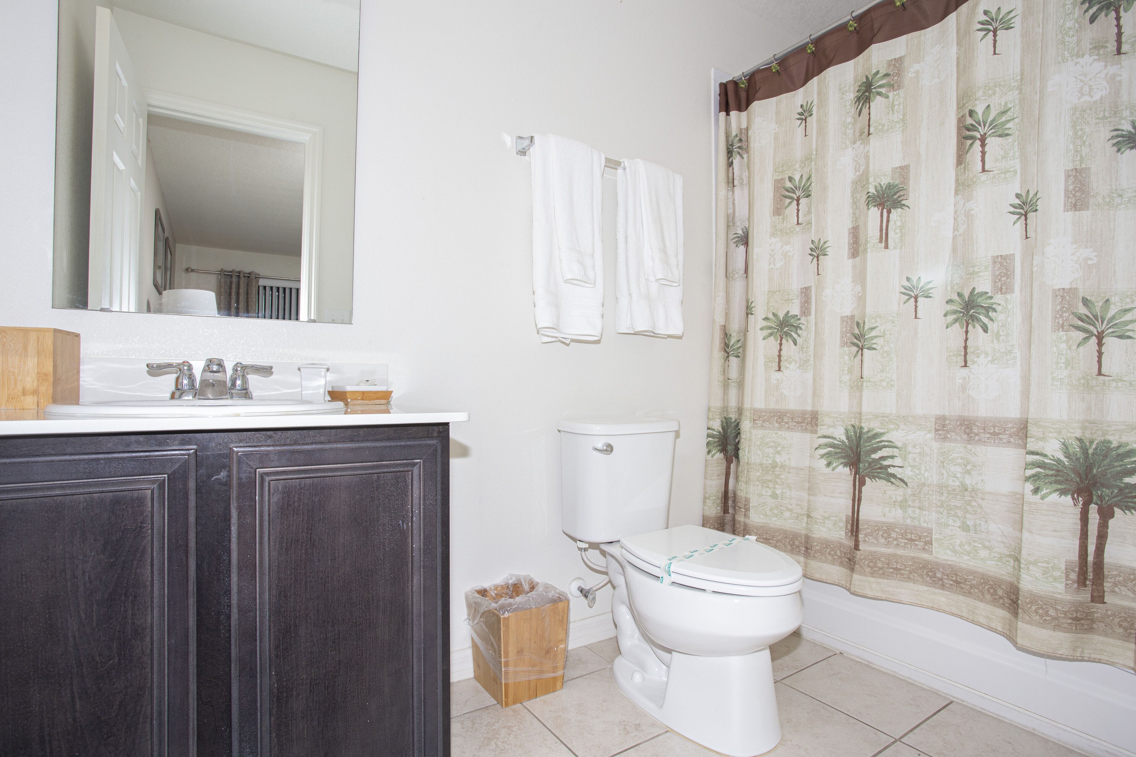 A bathroom with a toilet, sink, mirror, and a shower curtain with a palm tree pattern.