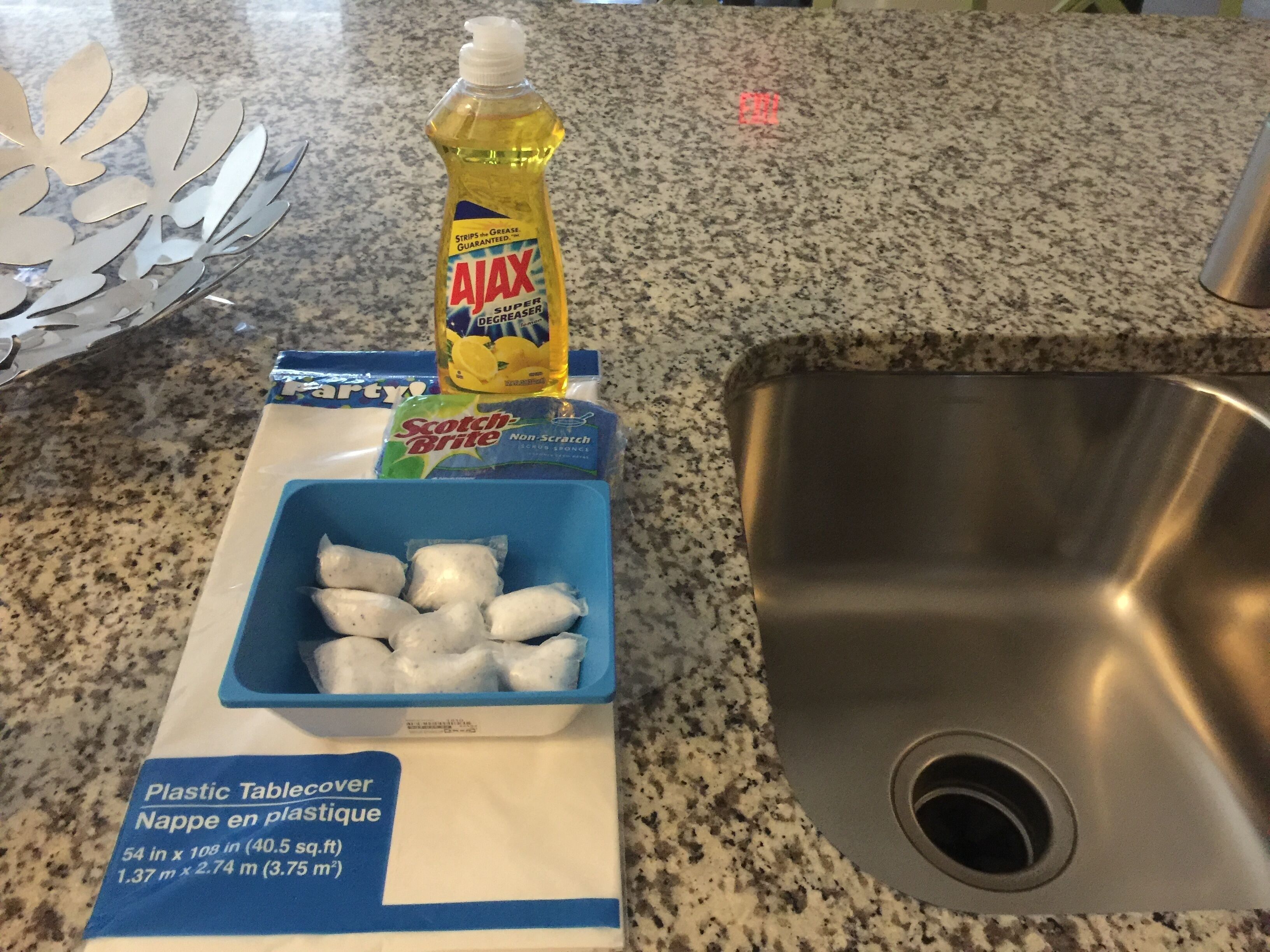 A kitchen counter with a stainless steel sink, a plastic tablecover, Ajax dish soap, Scotch-Brite non-scratch pads, and a blue container with white objects.