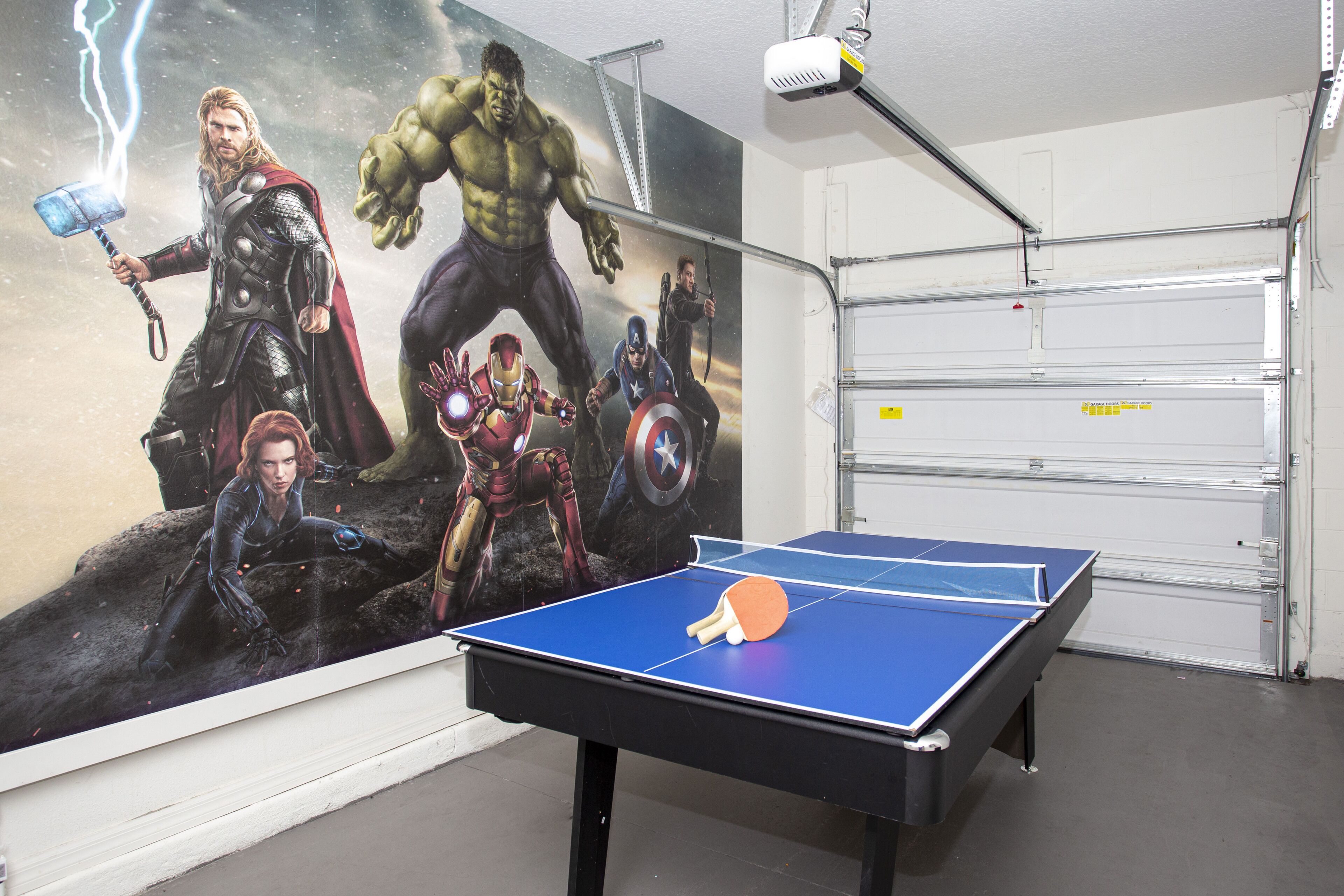 Un tavolo da ping pong in una stanza con un grande murale a tema supereroi sulla parete e un'ampia porta garage sullo sfondo.