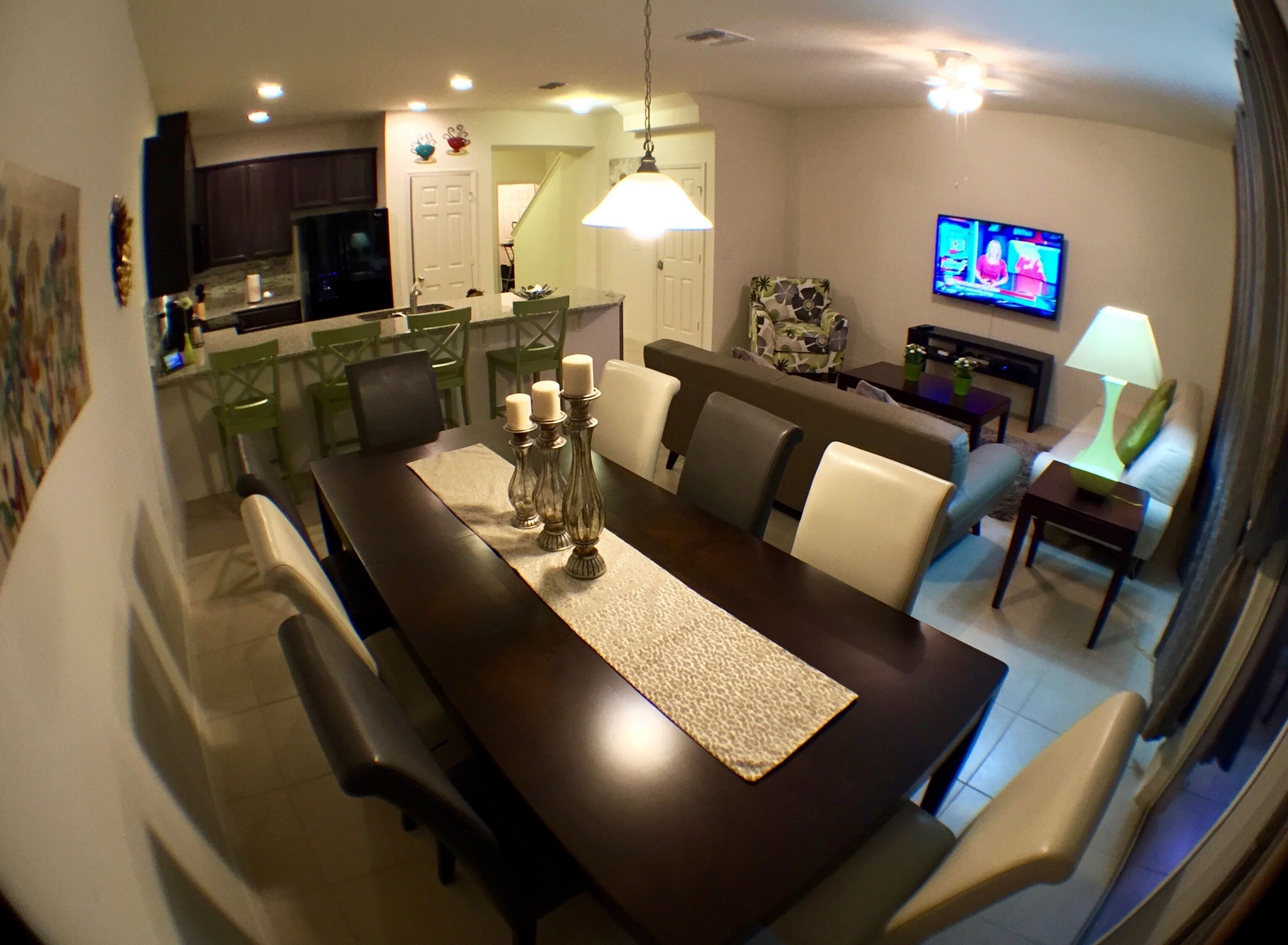 Un espace repas avec une longue table, des chaises, un lustre et un téléviseur en arrière-plan.