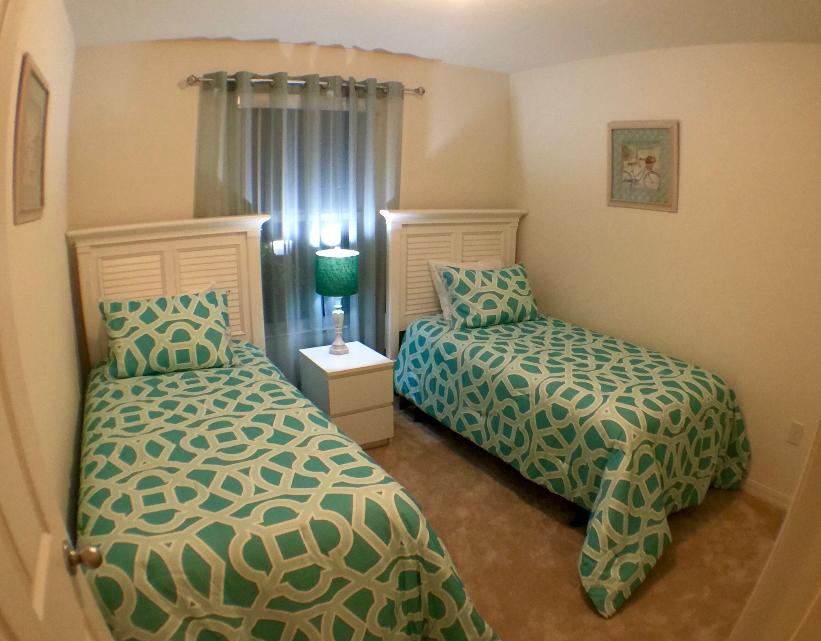 A bedroom with two single beds, a nightstand with a lamp, a window with curtains, and a framed picture on the wall.