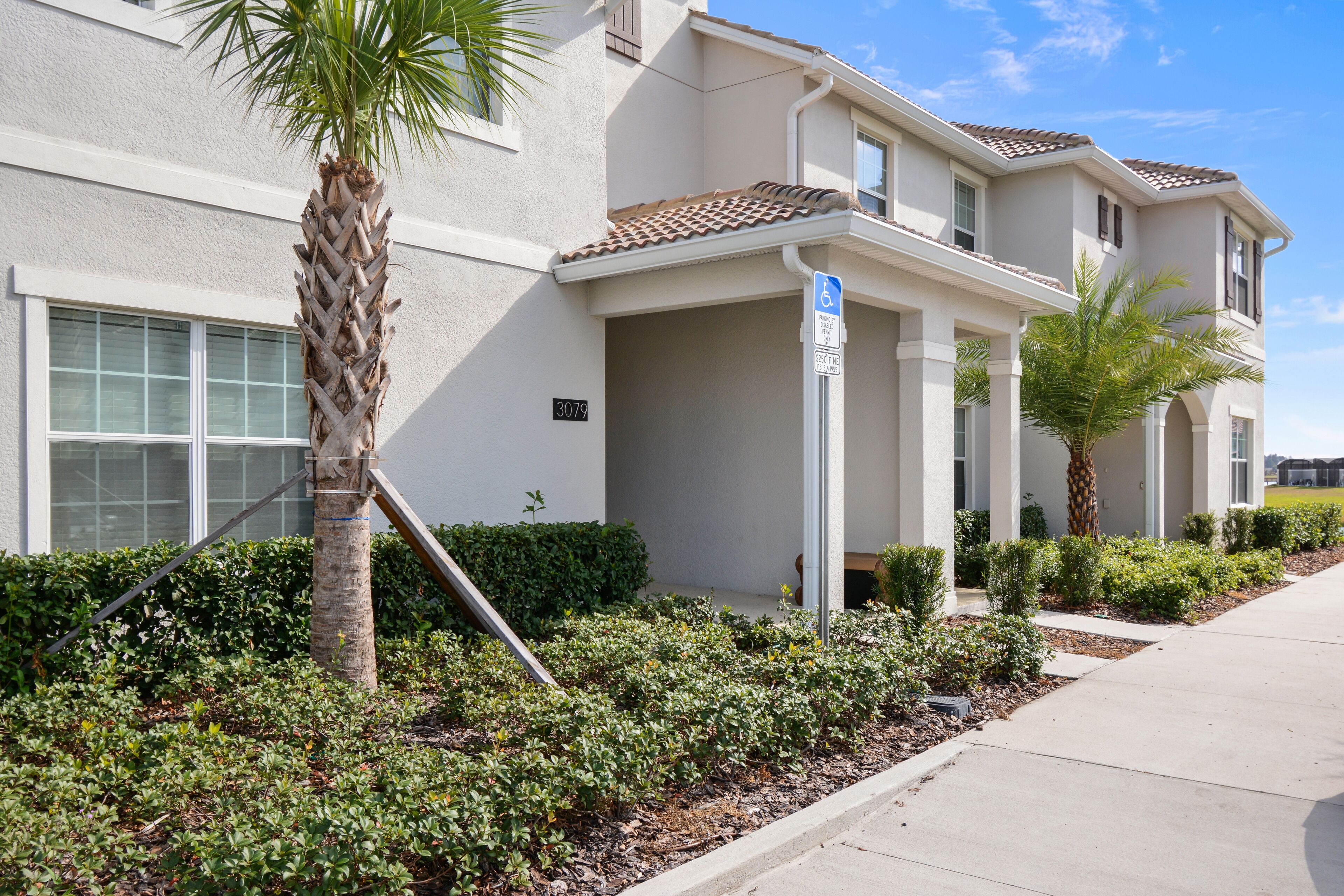 A multi-story residential building with a beige exterior, a tiled roof, and a well-maintained front yard with various plants and a palm tree.
