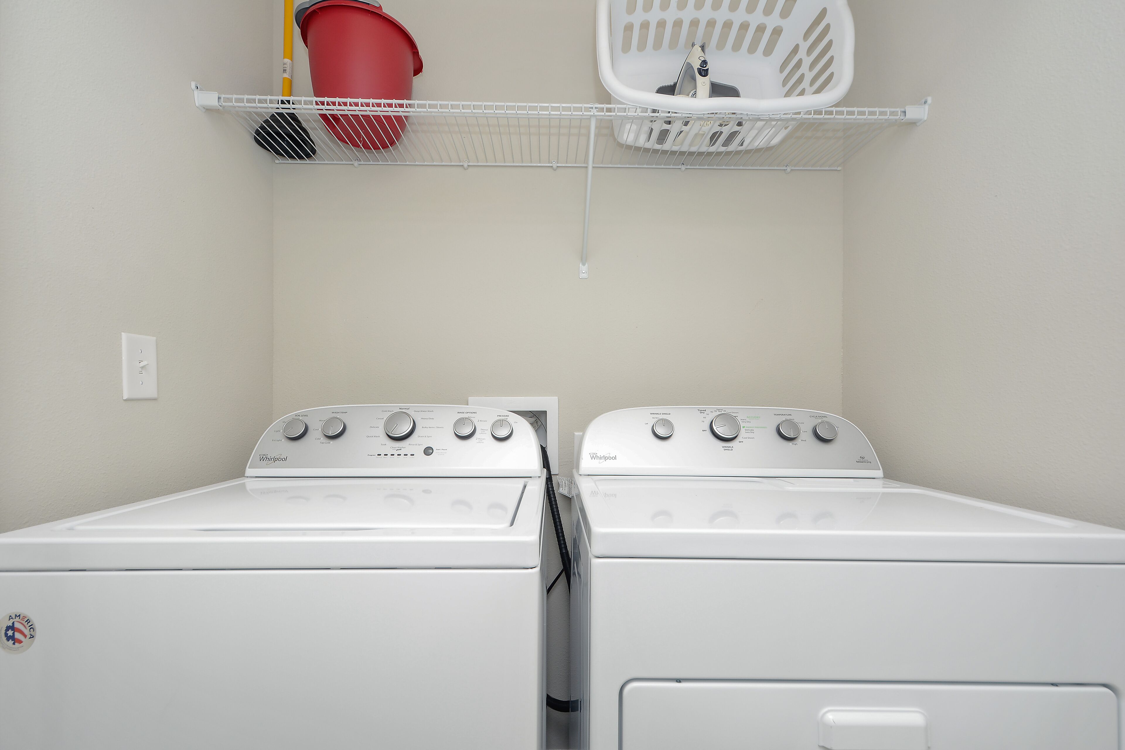 A laundry room with two white Whirlpool washers and dryers, a red bucket, and a white laundry basket on a shelf.
