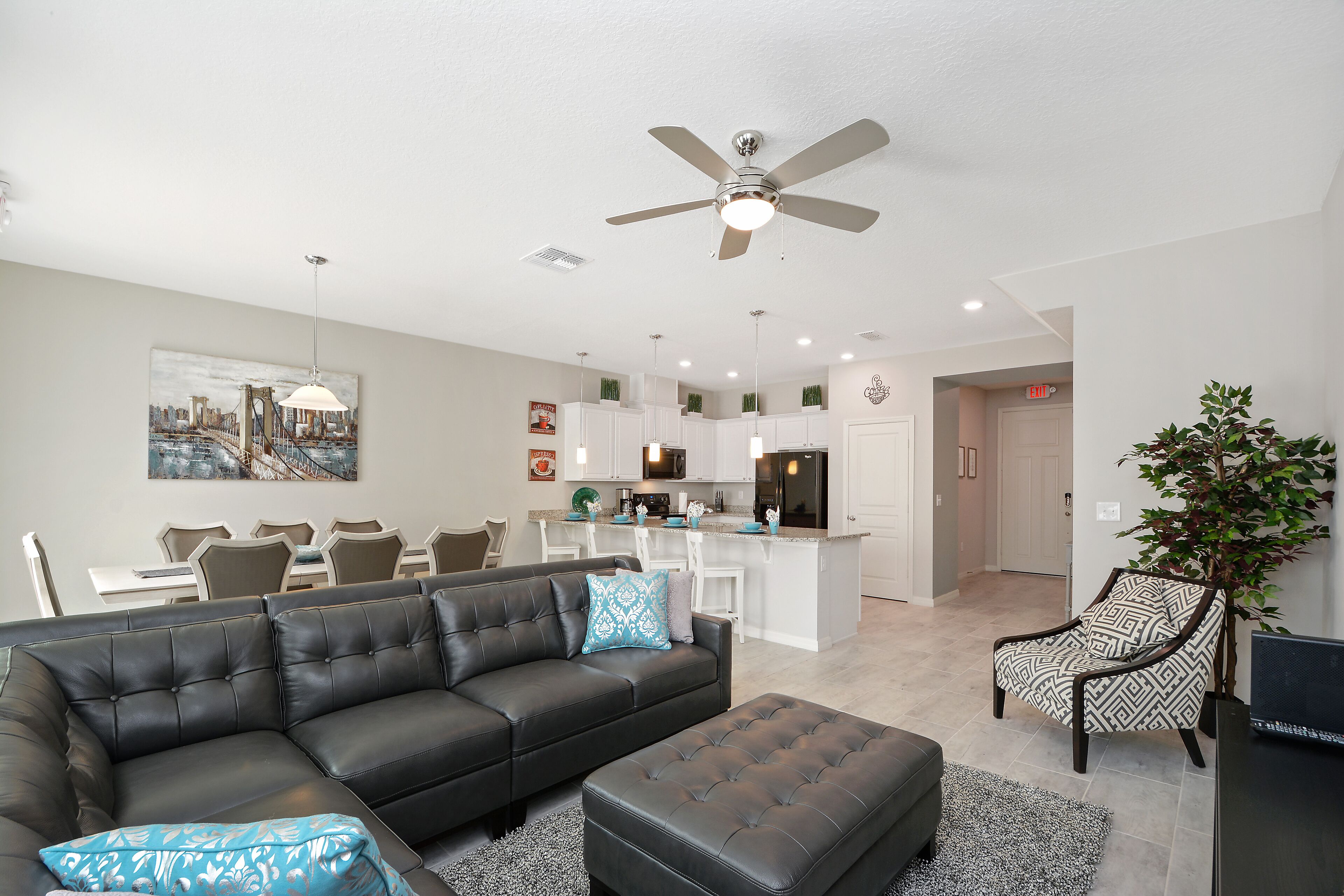 A modern living room with a black leather sofa, a matching ottoman, and a patterned armchair. A ceiling fan and recessed lighting are visible.