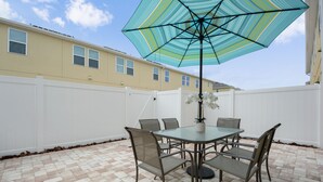 A patio with a glass table and chairs, a striped umbrella, and a white fence. - Family Friendly 4 Bedroom close to Disney in Compass Bay Resort 5108 (Kissimmee)