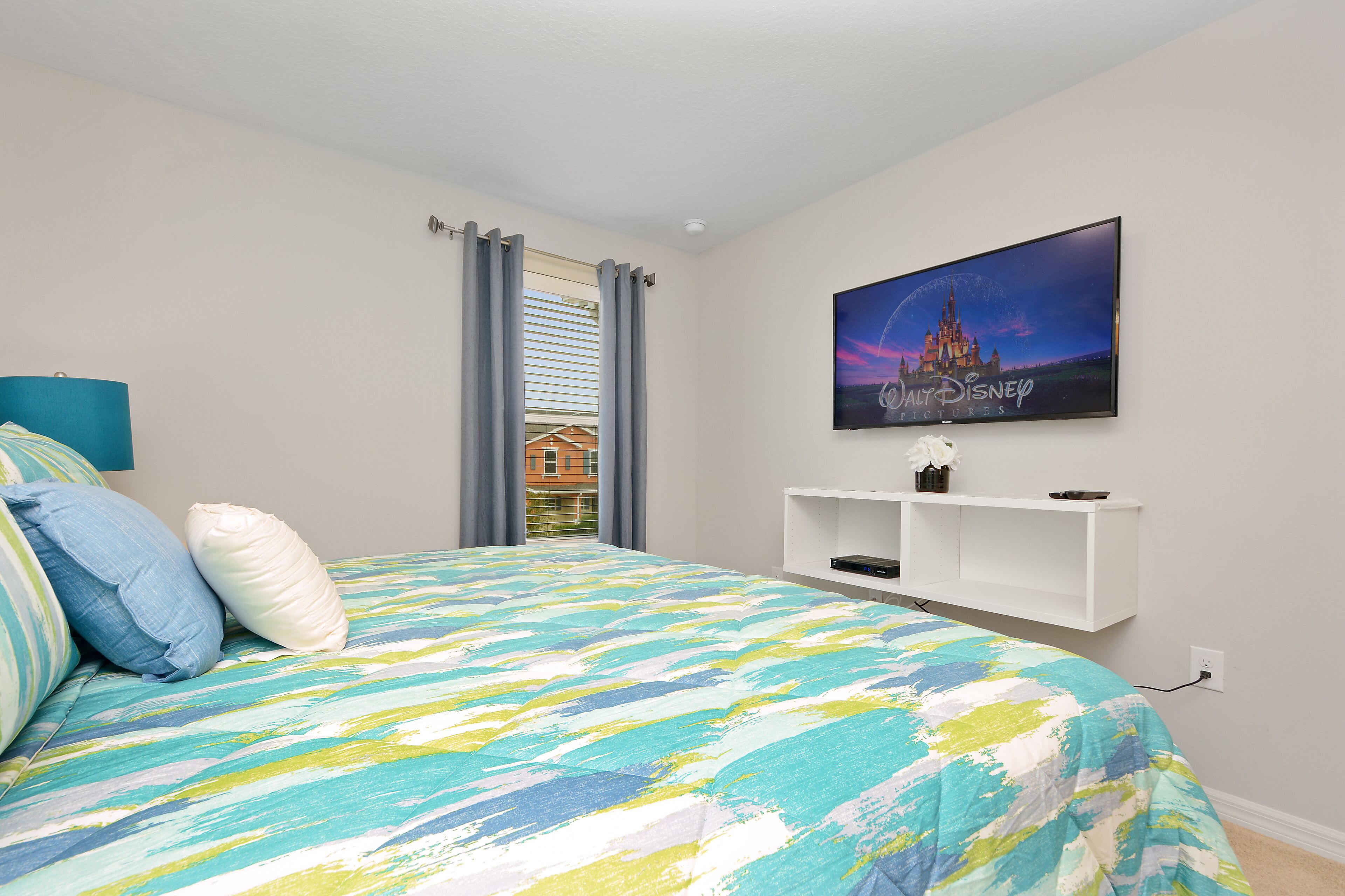 Ein Schlafzimmer mit einem Bett, einem an der Wand montierten Fernseher und einem Fenster mit Vorhängen.