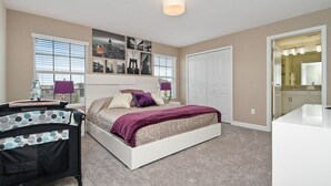A bedroom with a bed, a crib, a nightstand, a wall-mounted shelf with pictures, and a door leading to a bathroom.