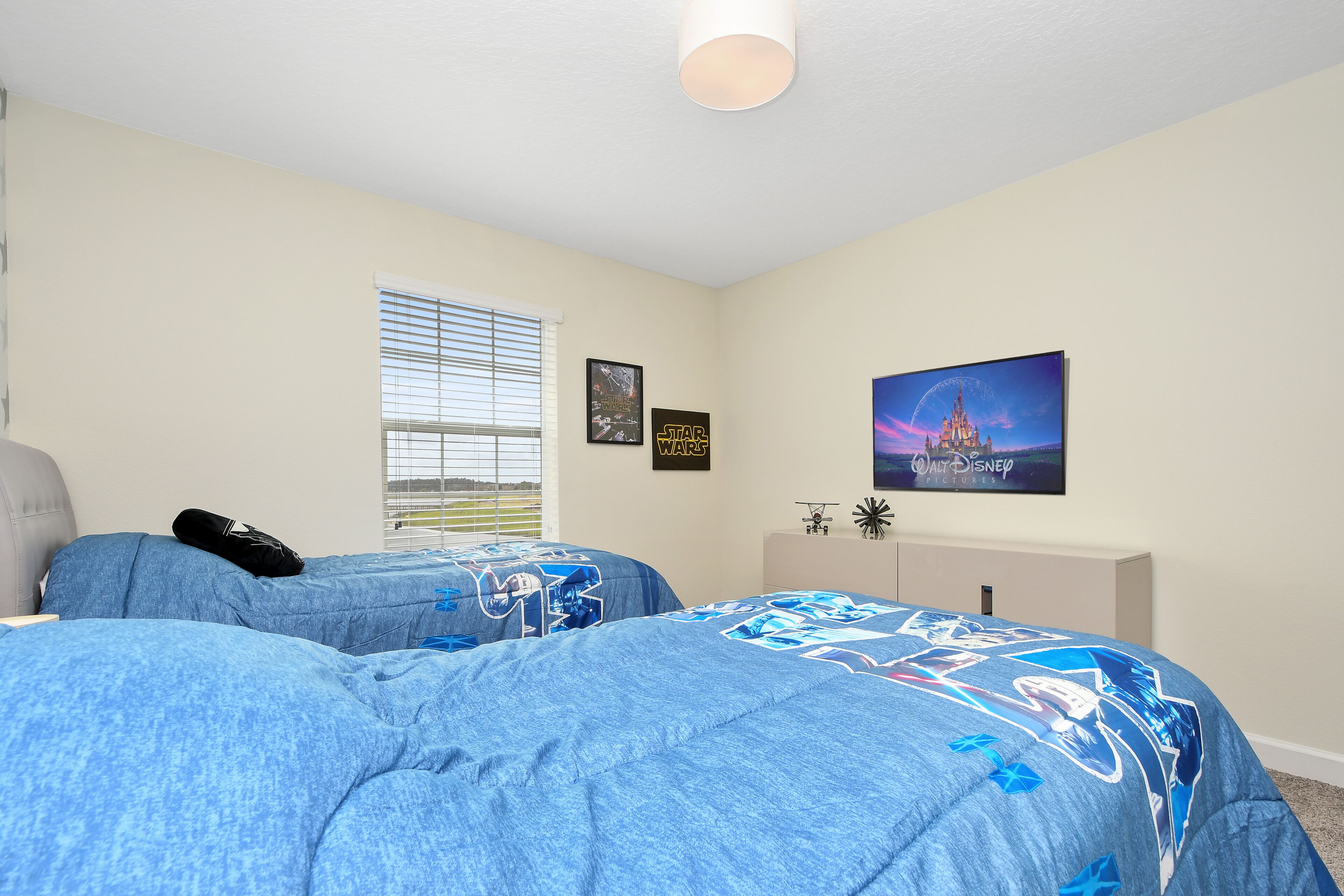 A bedroom with two beds, a window with blinds, a Star Wars poster, a Disney poster, and a small white cabinet.