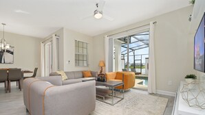 A modern living room with a grey sofa, orange armchair, and a coffee table. There is a dining area with chairs and a table, and a large window with a view of a pool.