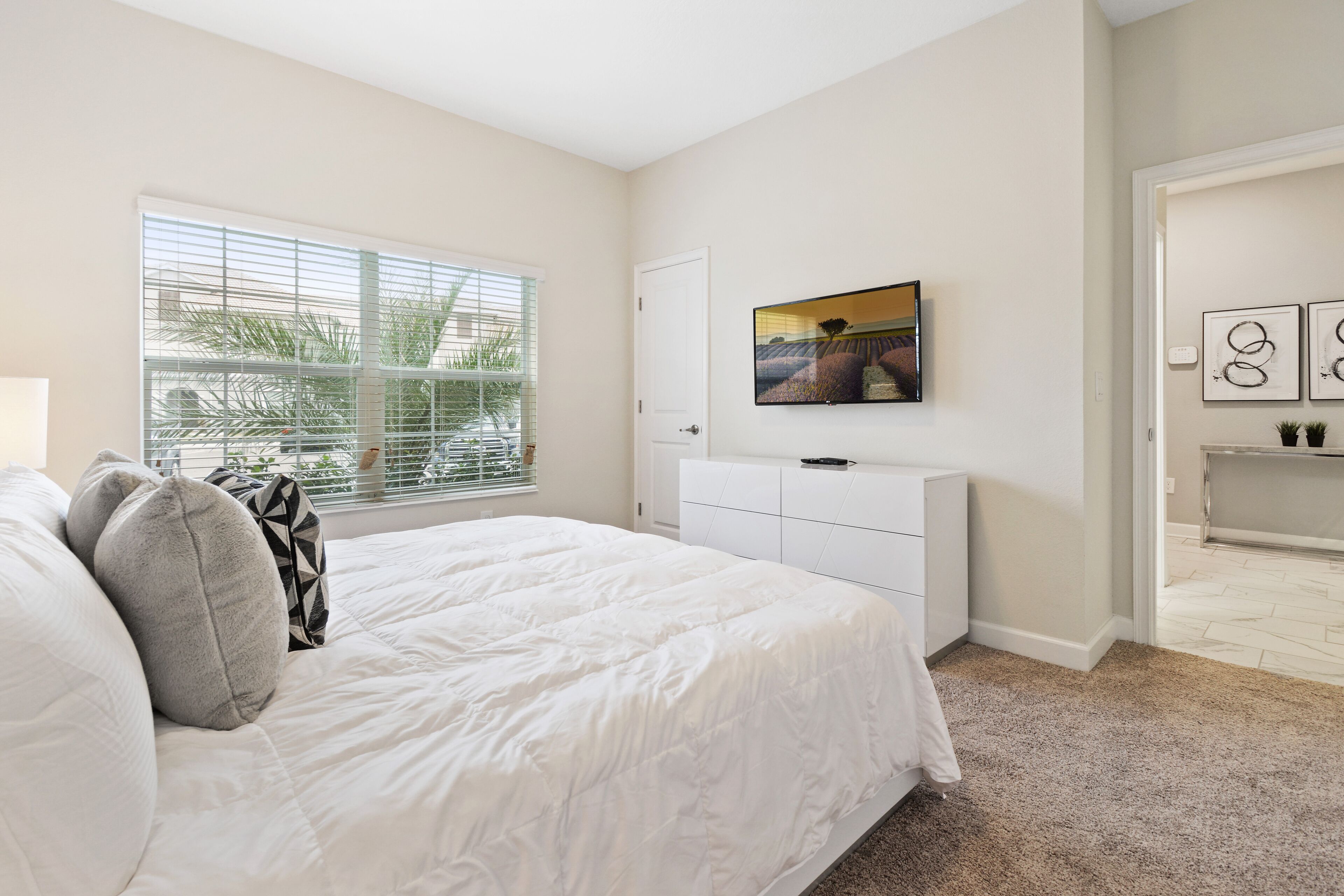 A bedroom with a large bed, a TV mounted on the wall, and a window with blinds.