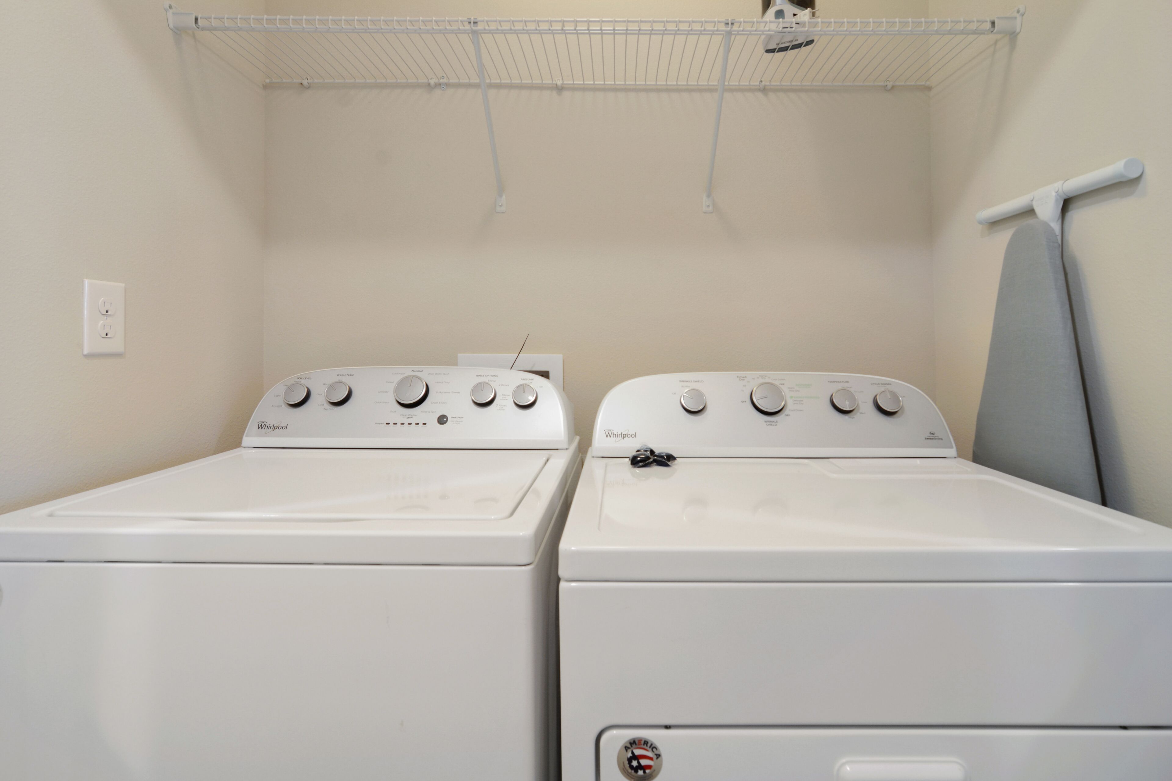 Two white Whirlpool dryers in a laundry room with a white ceiling and a metal shelf above.
