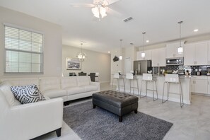 A modern living room with a white sofa, a grey ottoman, and a kitchen area with white cabinets and stainless steel appliances. - Family Friendly 4Bd PV Pool @ Storey L Resort 4896 (Kissimmee)