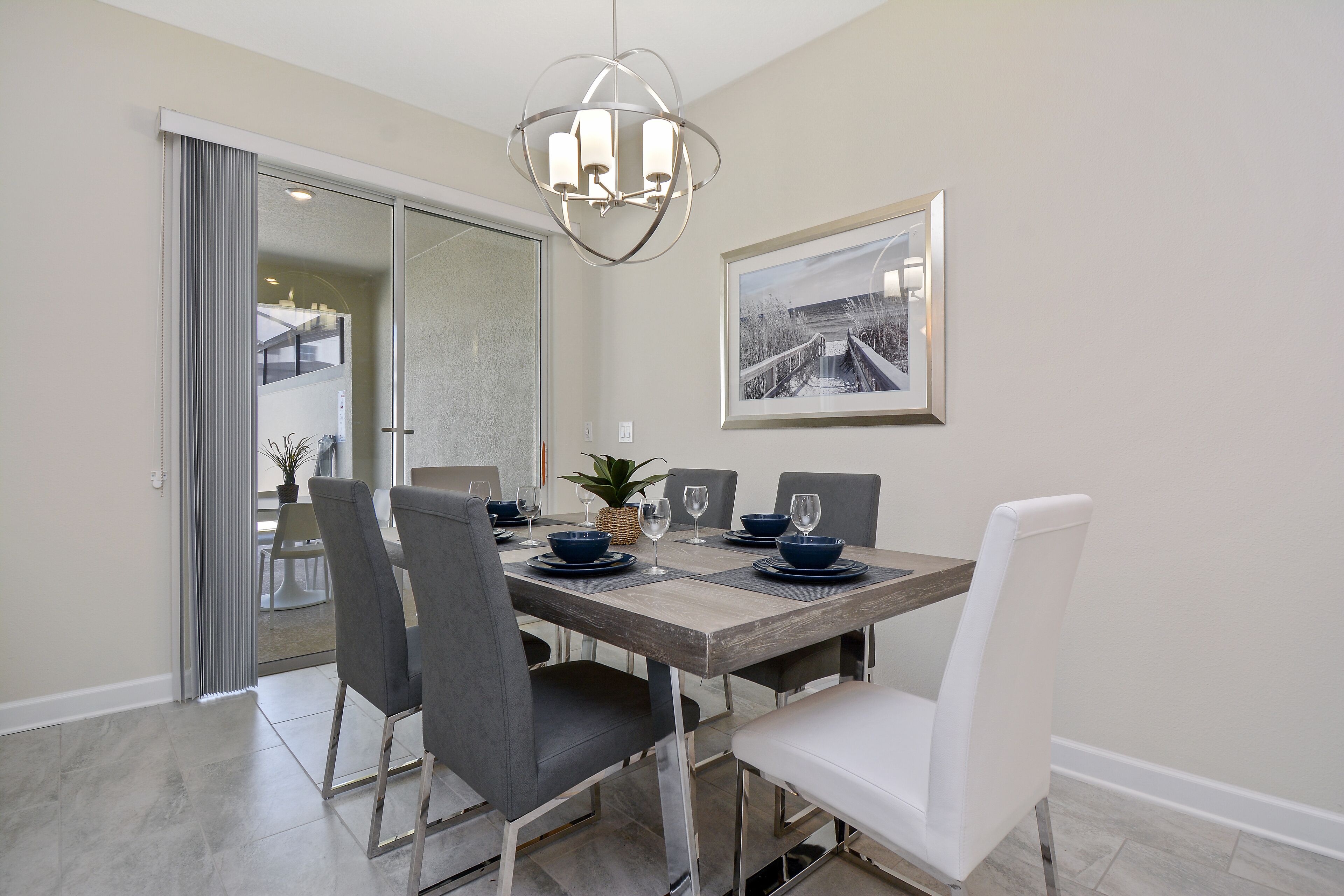 A dining area with a wooden table, grey and white chairs, a modern chandelier, and a framed picture on the wall.
