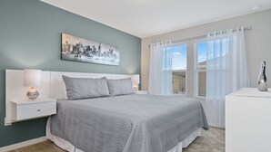 Une chambre à coucher avec un lit, des tables de chevet, une lampe, un tableau représentant un paysage urbain et une fenêtre avec des rideaux.