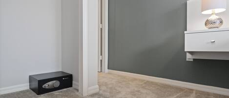 A room with a grey carpet, a white wall, a white nightstand with a lamp, and a black safe.