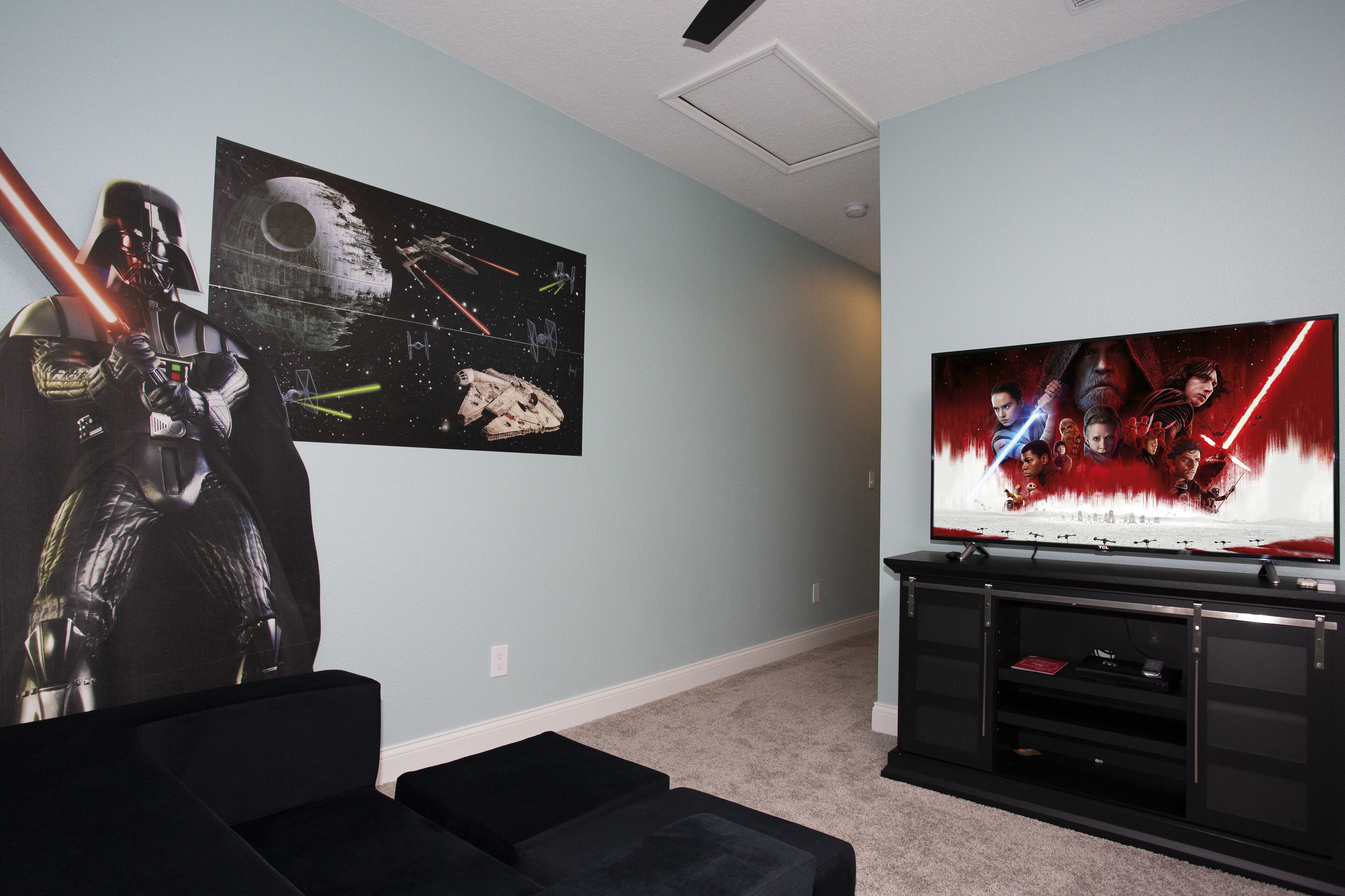 Una habitación con un televisor de pantalla plana grande montado en la pared, un mueble de entretenimiento negro debajo y carteles de Star Wars en la pared.