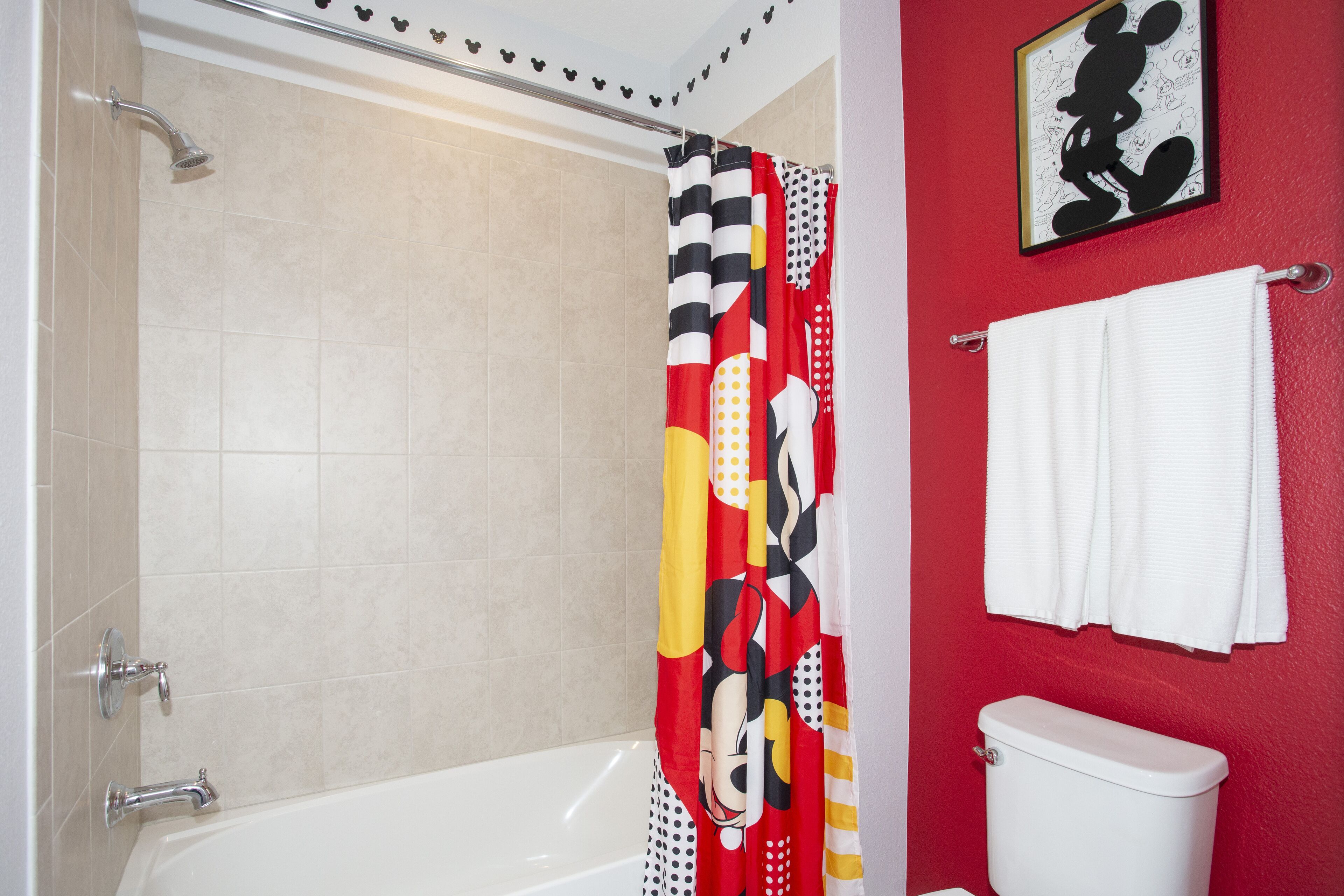 A bathroom with a shower curtain featuring a cartoon character, a white toilet, and a red wall with a Mickey Mouse framed picture.