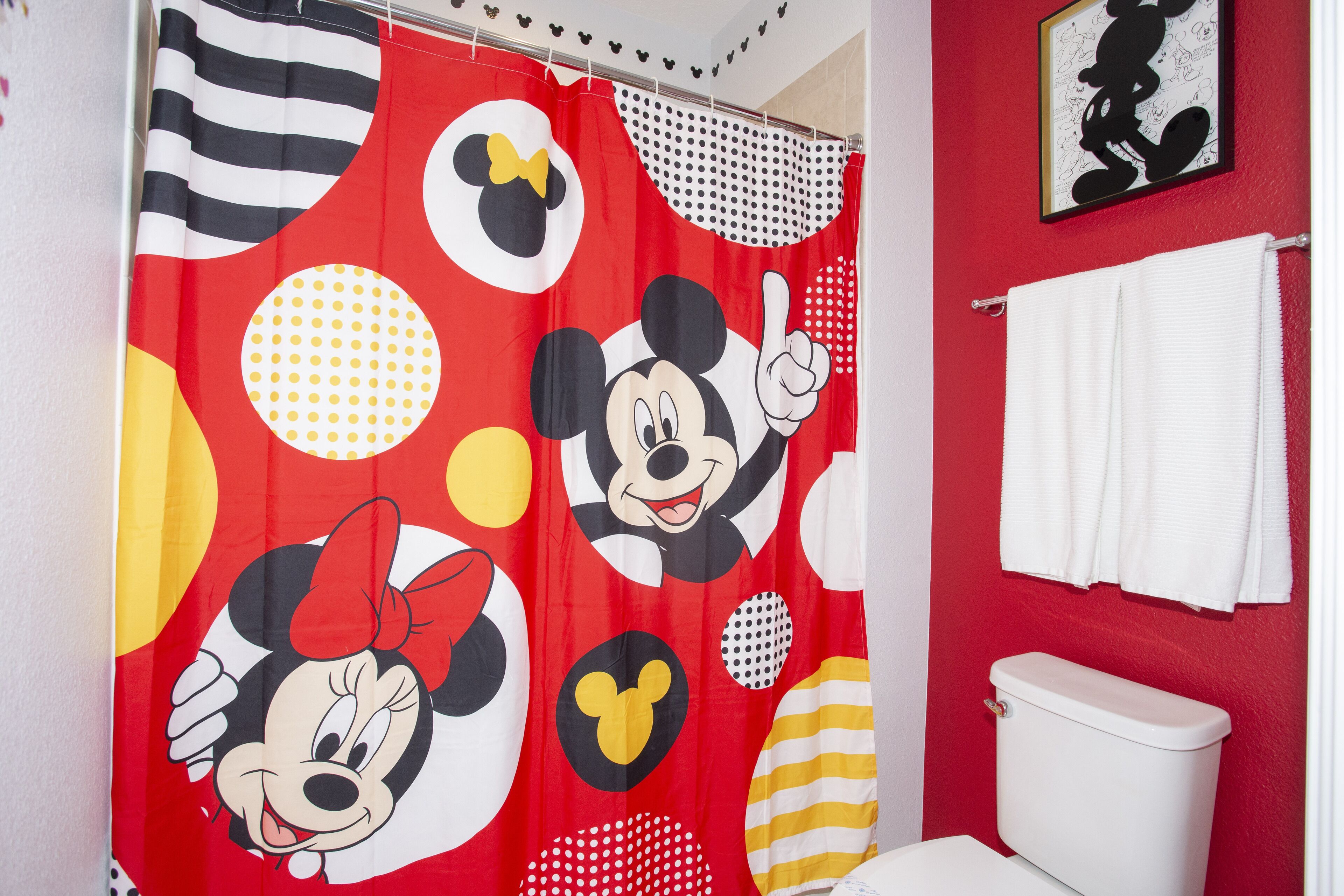 Un baño con cortina de ducha de Mickey Mouse, un inodoro blanco y una pared roja.