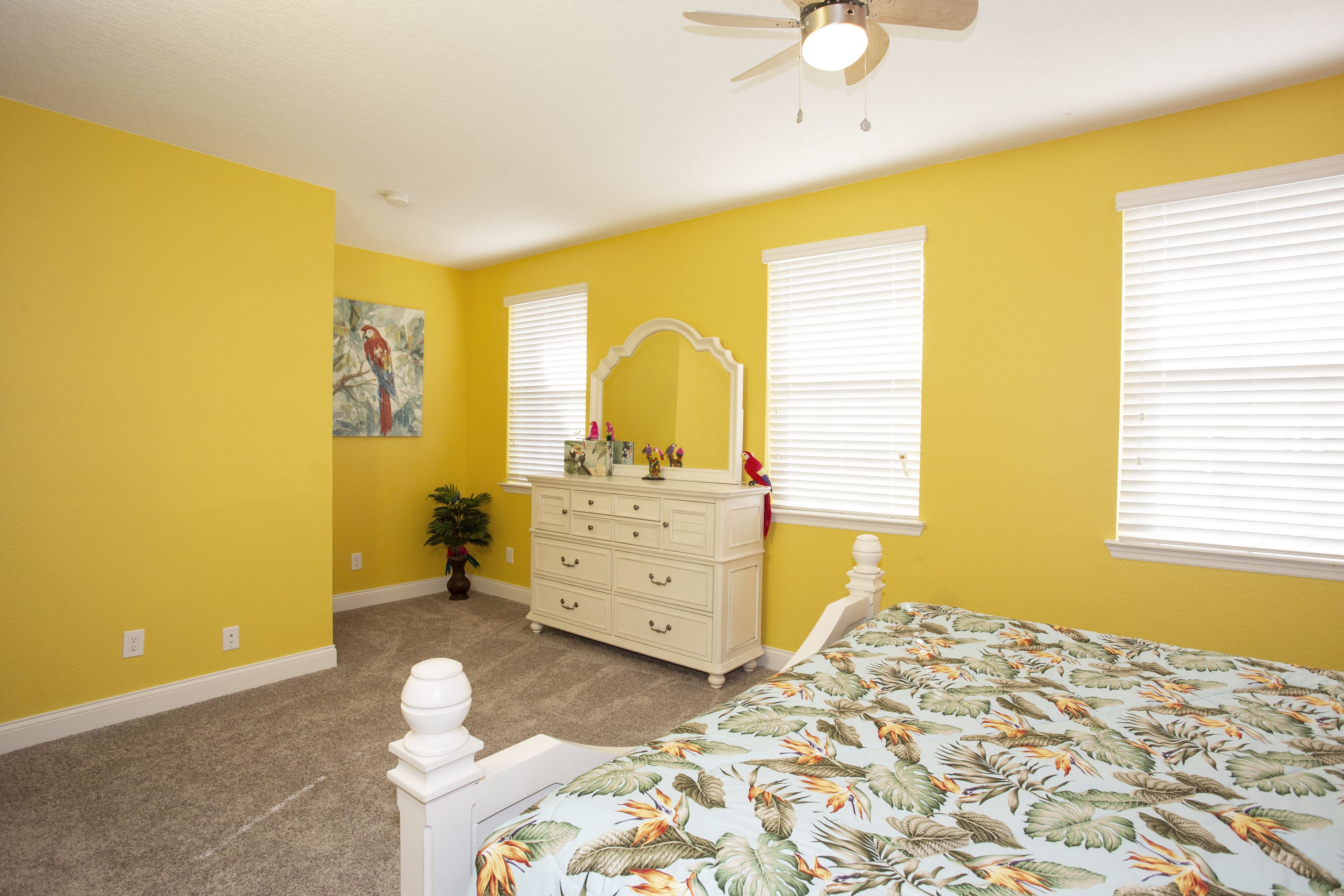 A bedroom with yellow walls, a white dresser, a bed with a tropical print bedspread, and a ceiling fan.