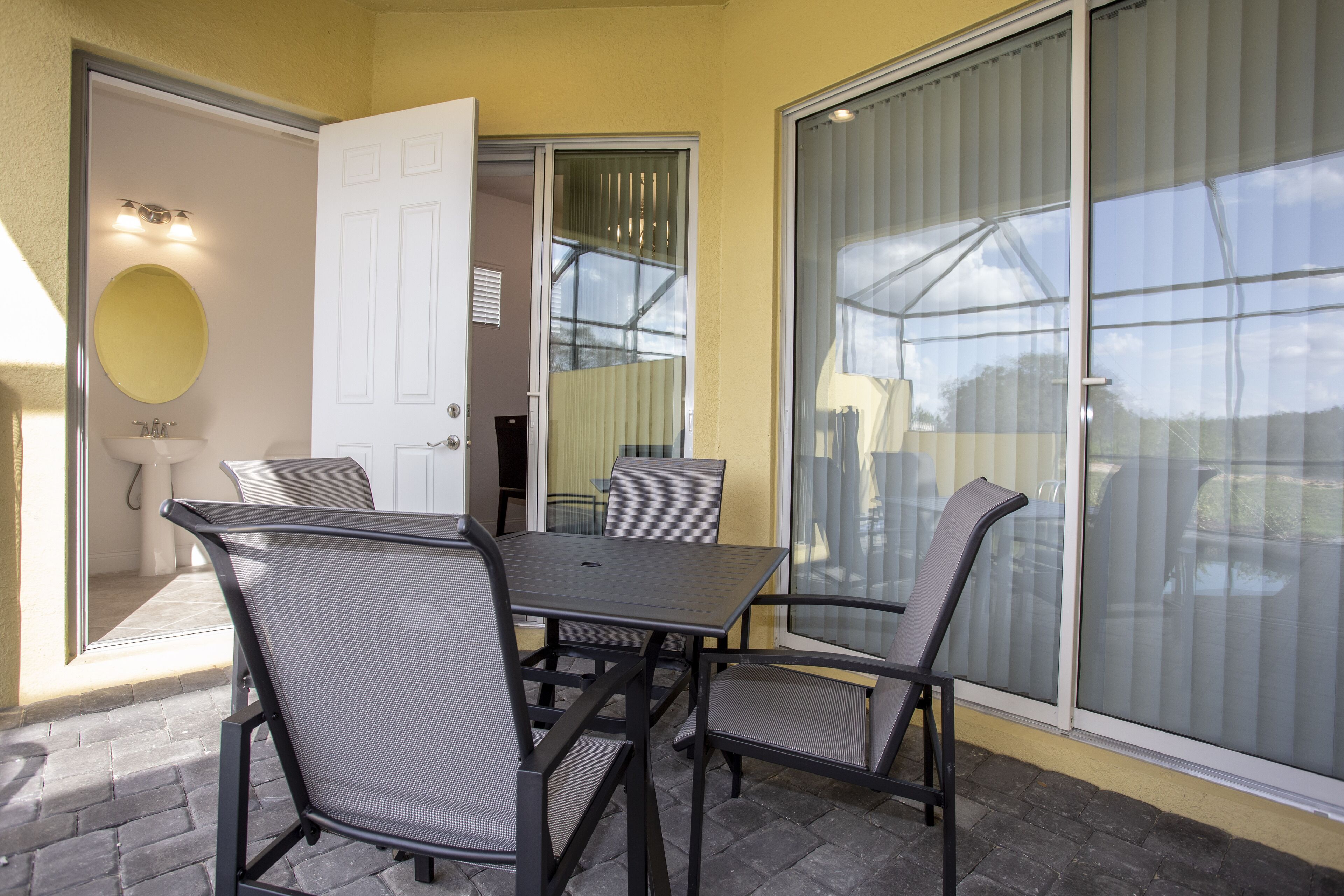 Un patio con una mesa y sillas, una puerta que da a un baño y un ventanal con persianas.