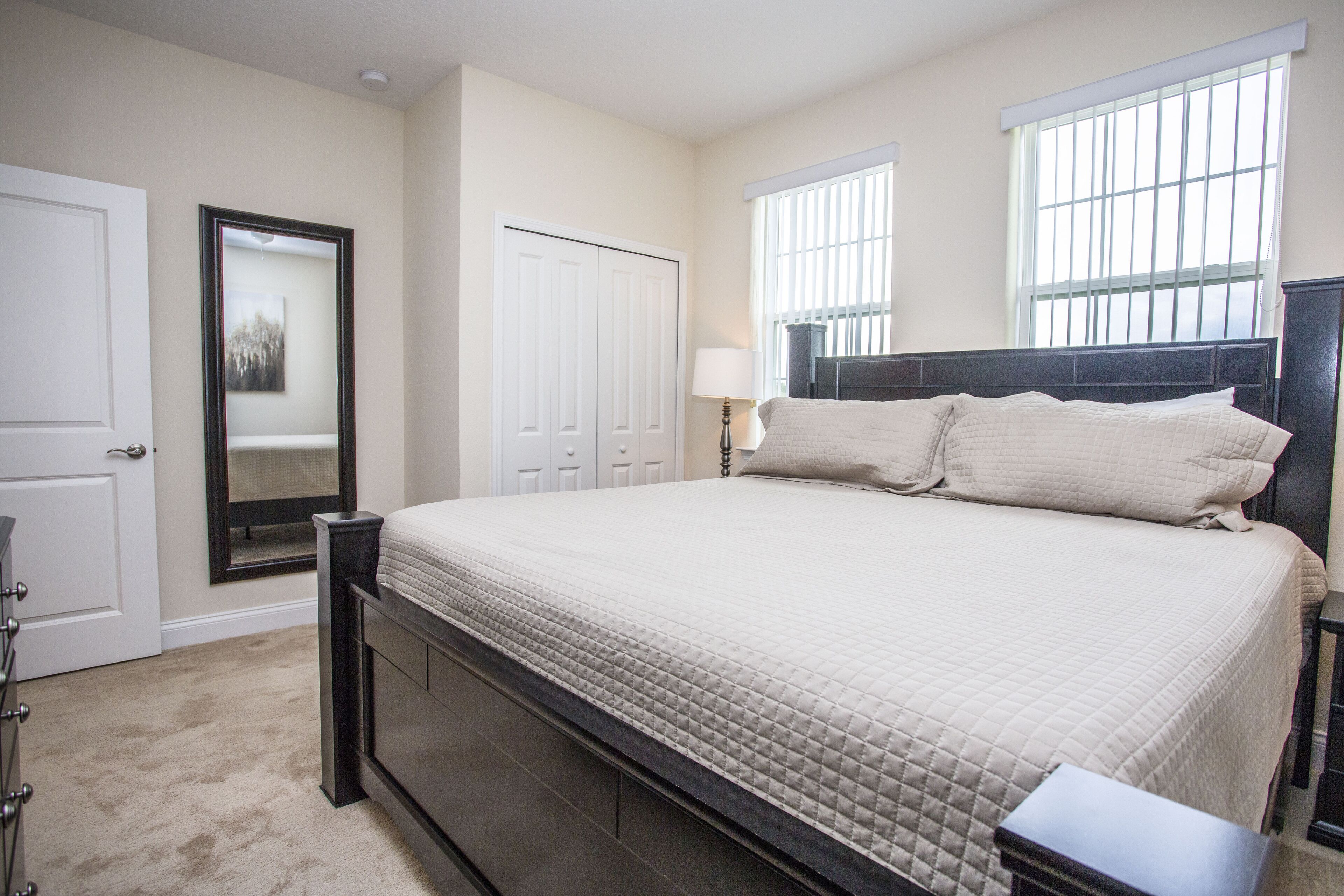 A bedroom with a large bed, a dresser, a mirror, and two windows.