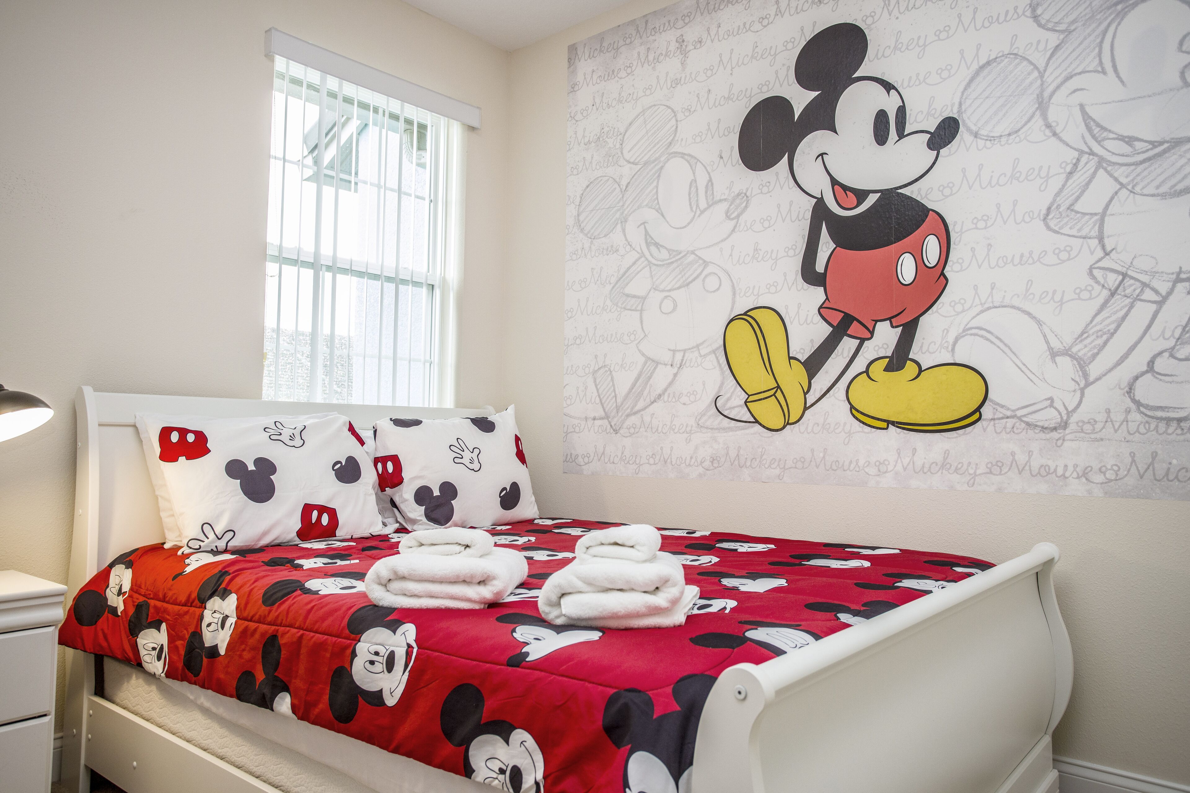 A hotel room with a Mickey Mouse themed bedspread, pillows, and wall art.
