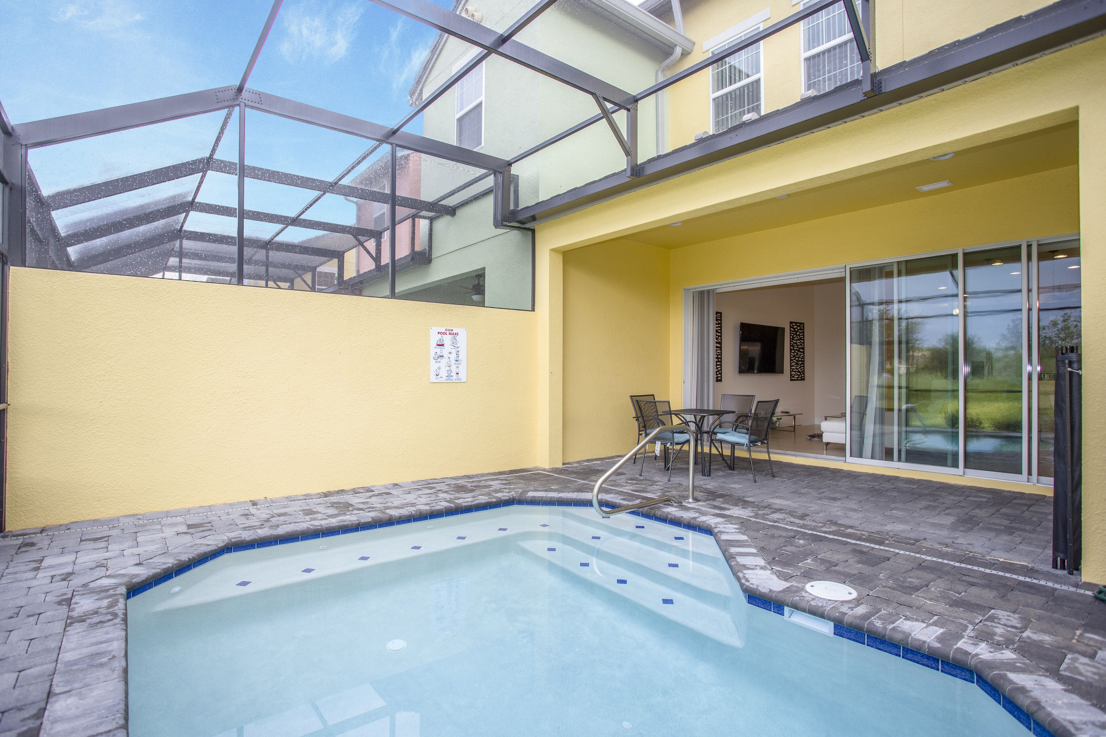 A swimming pool with a glass ceiling, a patio area with chairs, and a yellow building facade.