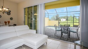 Un salon moderne avec un canapé blanc, un lustre et une vue sur une terrasse extérieure dotée d’une piscine et de chaises.