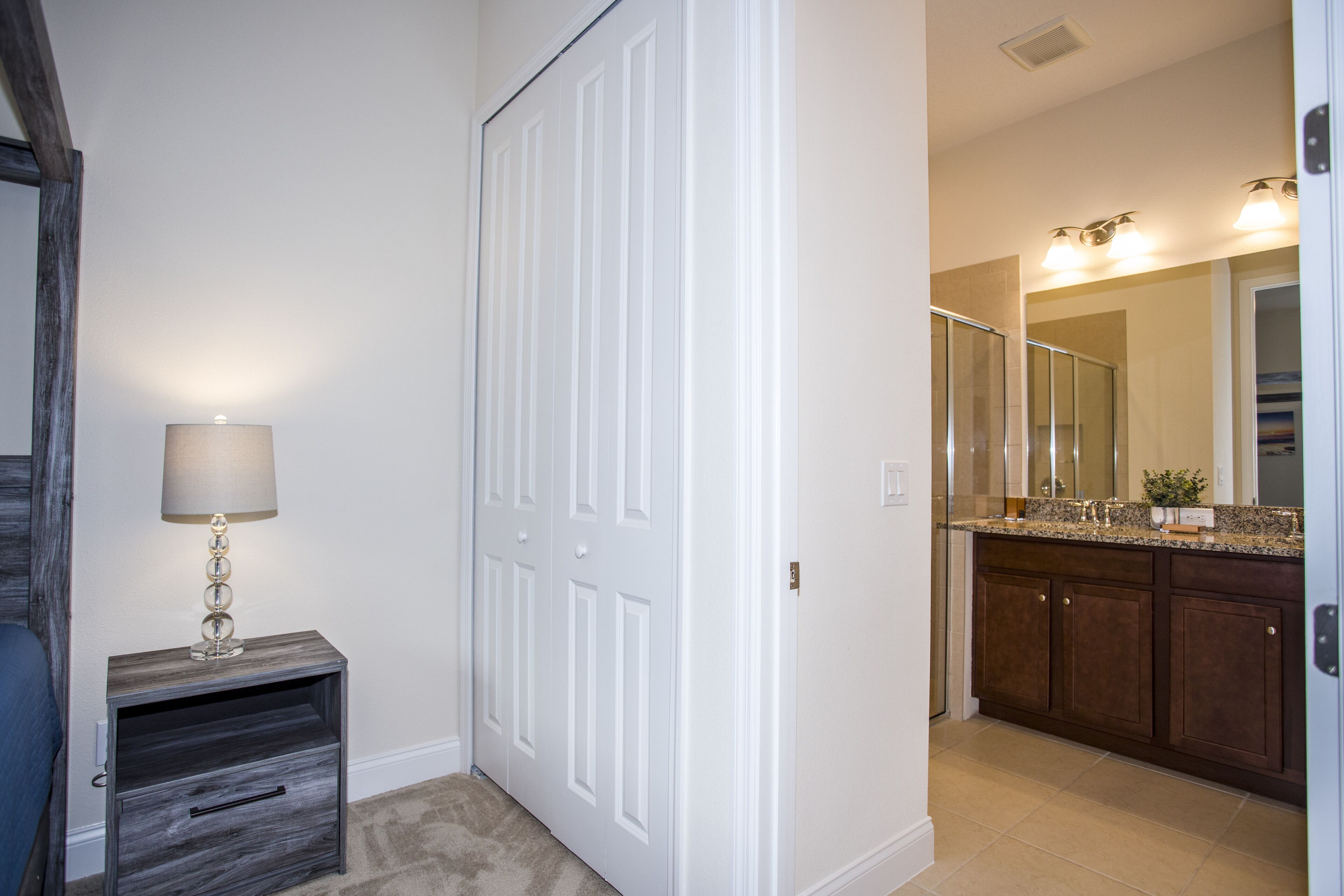 Un couloir avec une porte blanche fermée, une table de chevet avec une lampe, et une salle de bain avec un meuble-lavabo et un miroir.