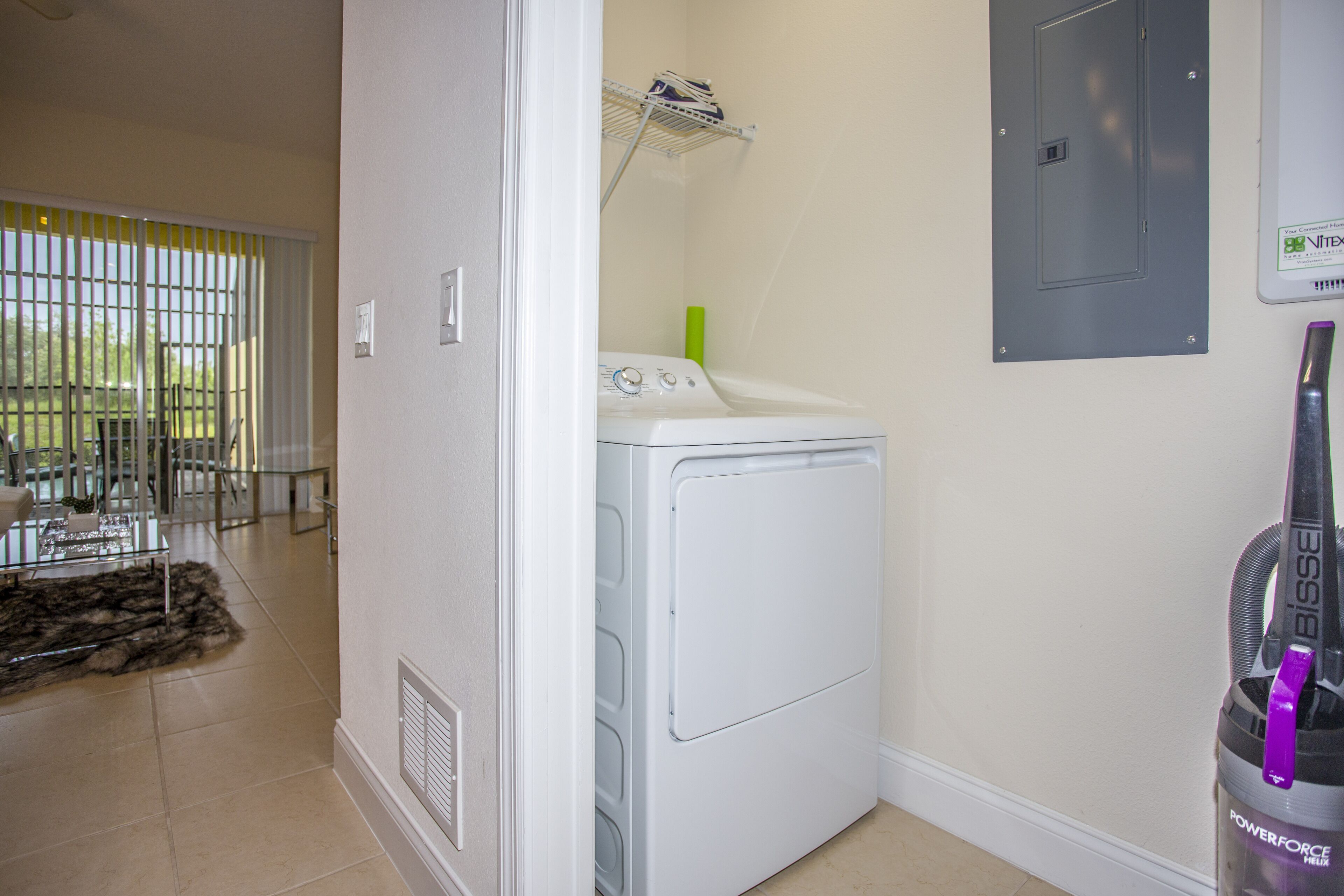 A laundry room with a front-loading washer and dryer, a metal shelf above the dryer, and a vacuum cleaner to the right.