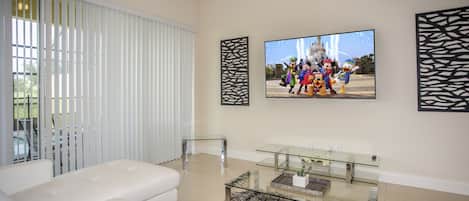 A modern living room with a white sofa, a glass coffee table, and a large flat-screen TV mounted on the wall.