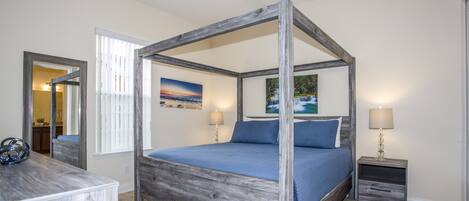 Un lit à baldaquin avec un cadre en bois, une couvre-lit bleue et des coussins assortis. Il y a deux tables de chevet avec des lampes, un grand miroir et, par une porte ouverte, on peut voir une cuisine et une salle de bain.