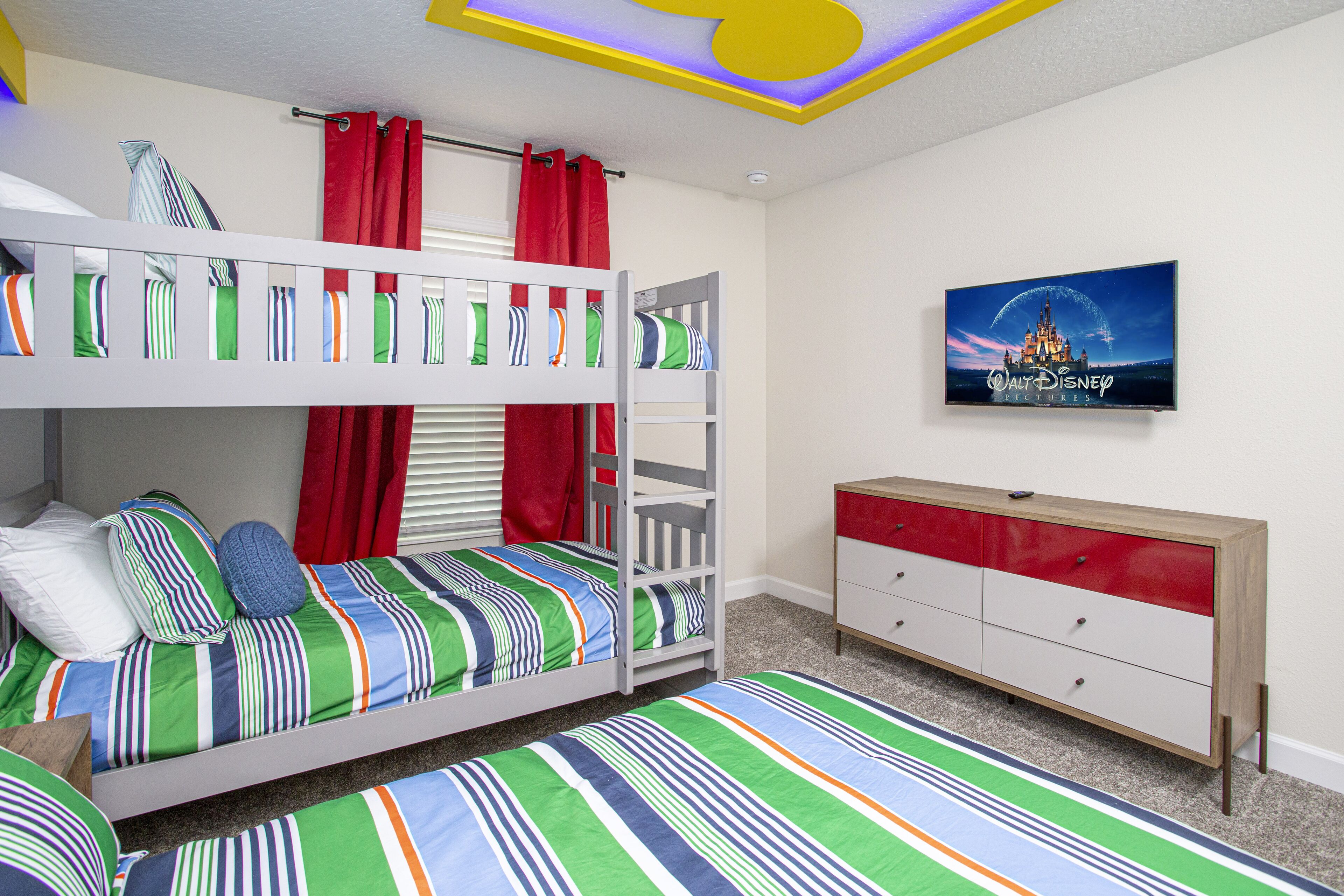 Ein Stockbett mit gestreifter Bettwäsche und roten Vorhängen, eine Kommode mit roten und weißen Schubladen und ein Disney-Poster an der Wand.