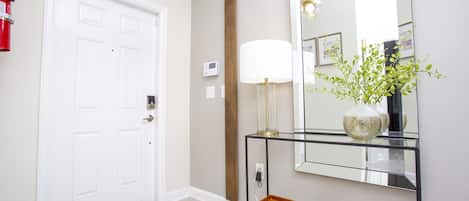 Un pasillo con una puerta blanca, un escritorio de cristal con una lámpara, un banco y un espejo que refleja una planta y adornos de pared.