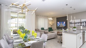Una cocina moderna con una isla central, un comedor con mesa de vidrio y una sala de estar con sofá y televisor.