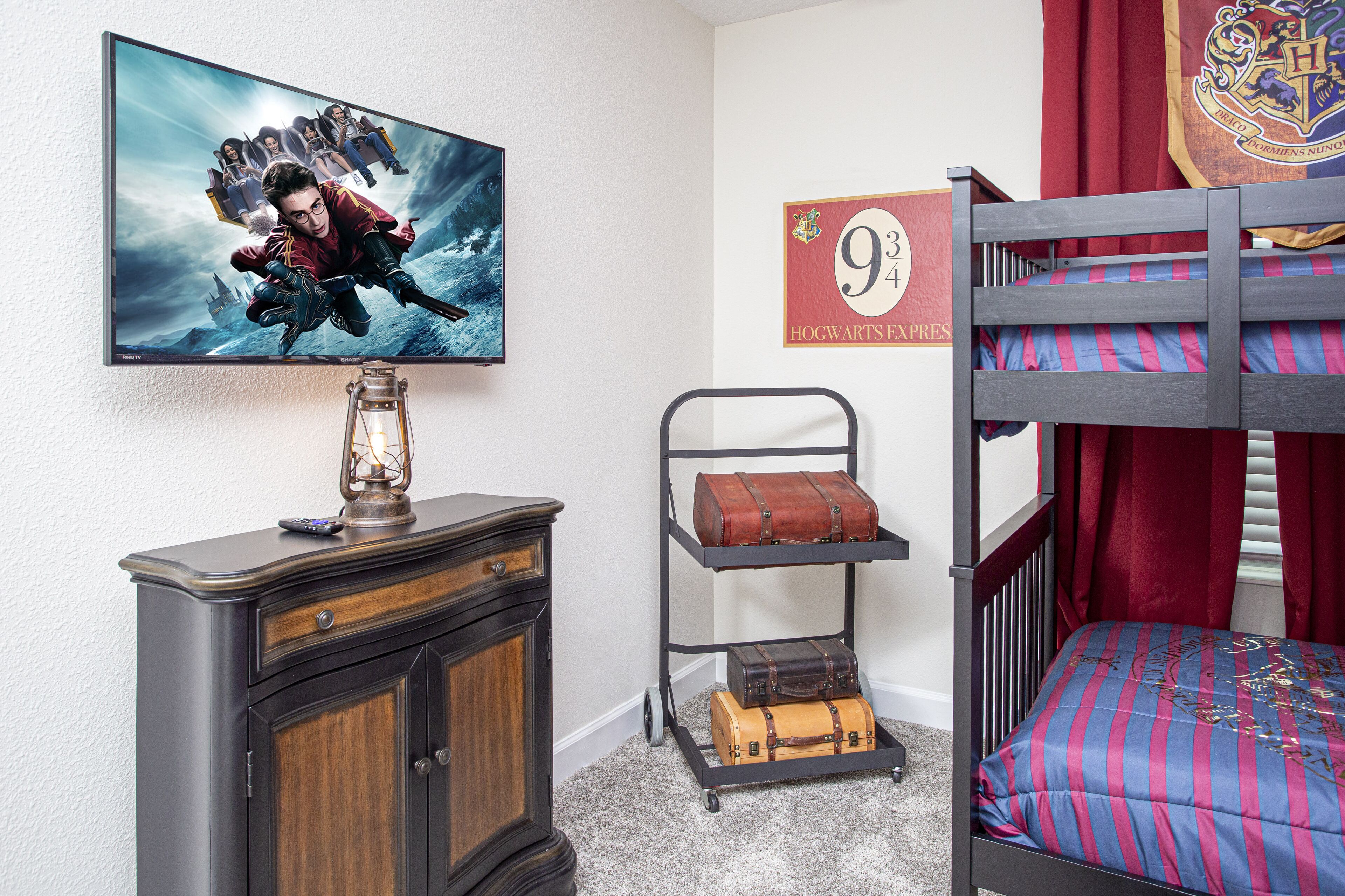 A room with a bunk bed, a dresser, a wall-mounted TV displaying a movie, a Hogwarts Express sign, and a luggage cart.
