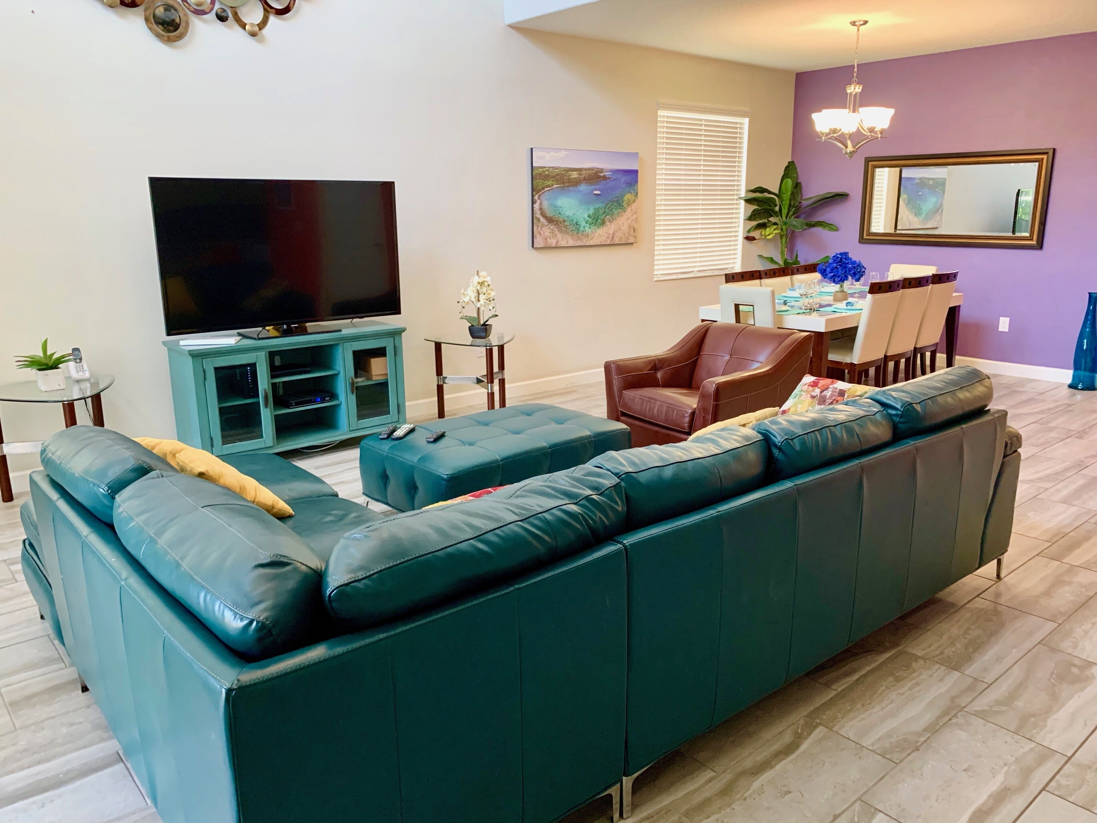 A living room with a teal sofa, a brown leather chair, a flat-screen TV, a dining table with chairs, and a potted plant.