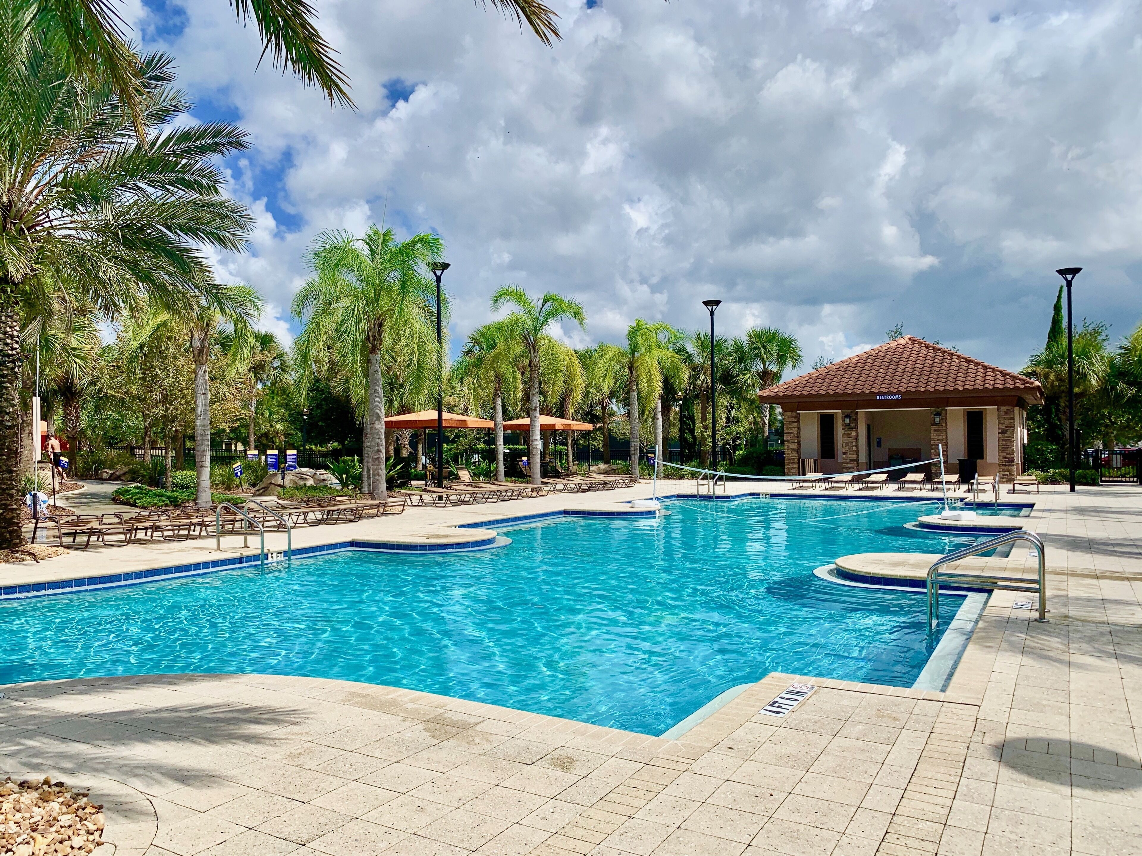 A swimming pool with lounge chairs and umbrellas, surrounded by palm trees and a building labeled "Restrooms".