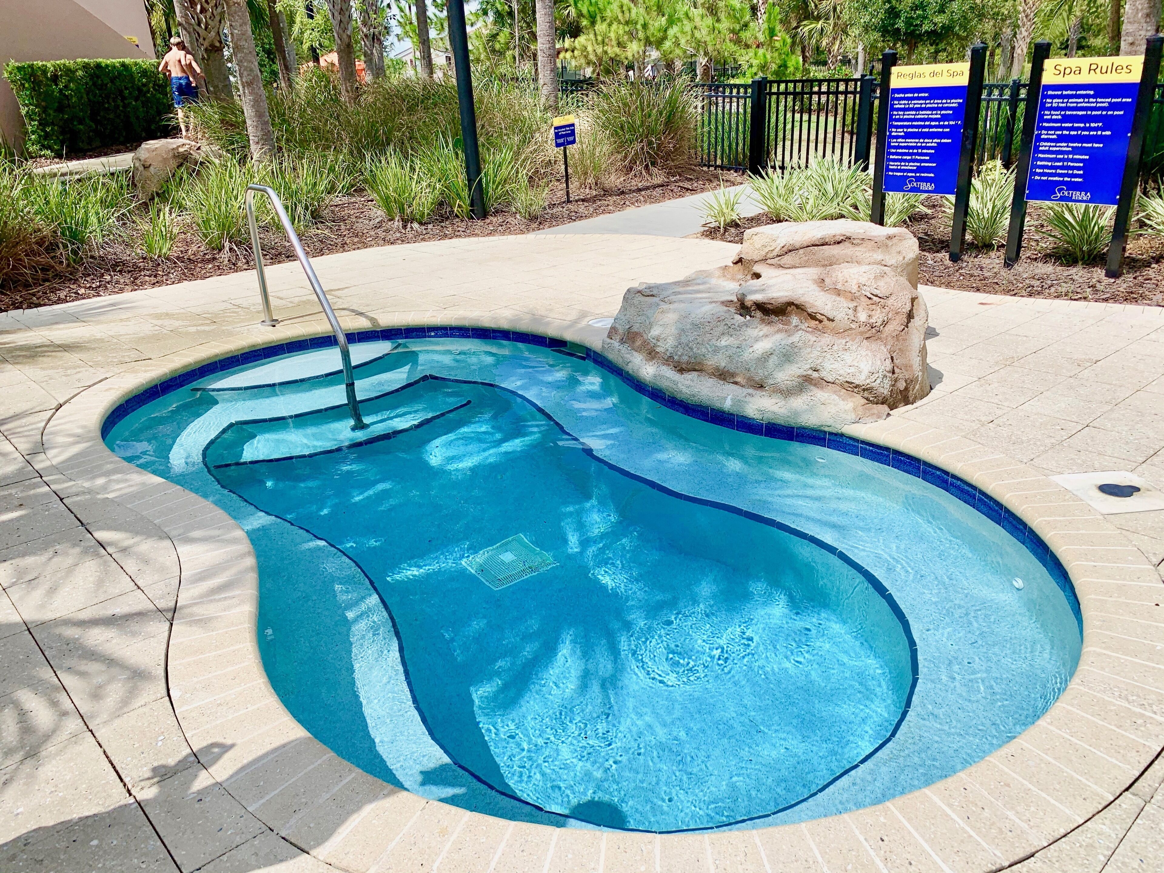 Una piscina con una roccia decorativa, circondata da una zona pavimentata e una recinzione.